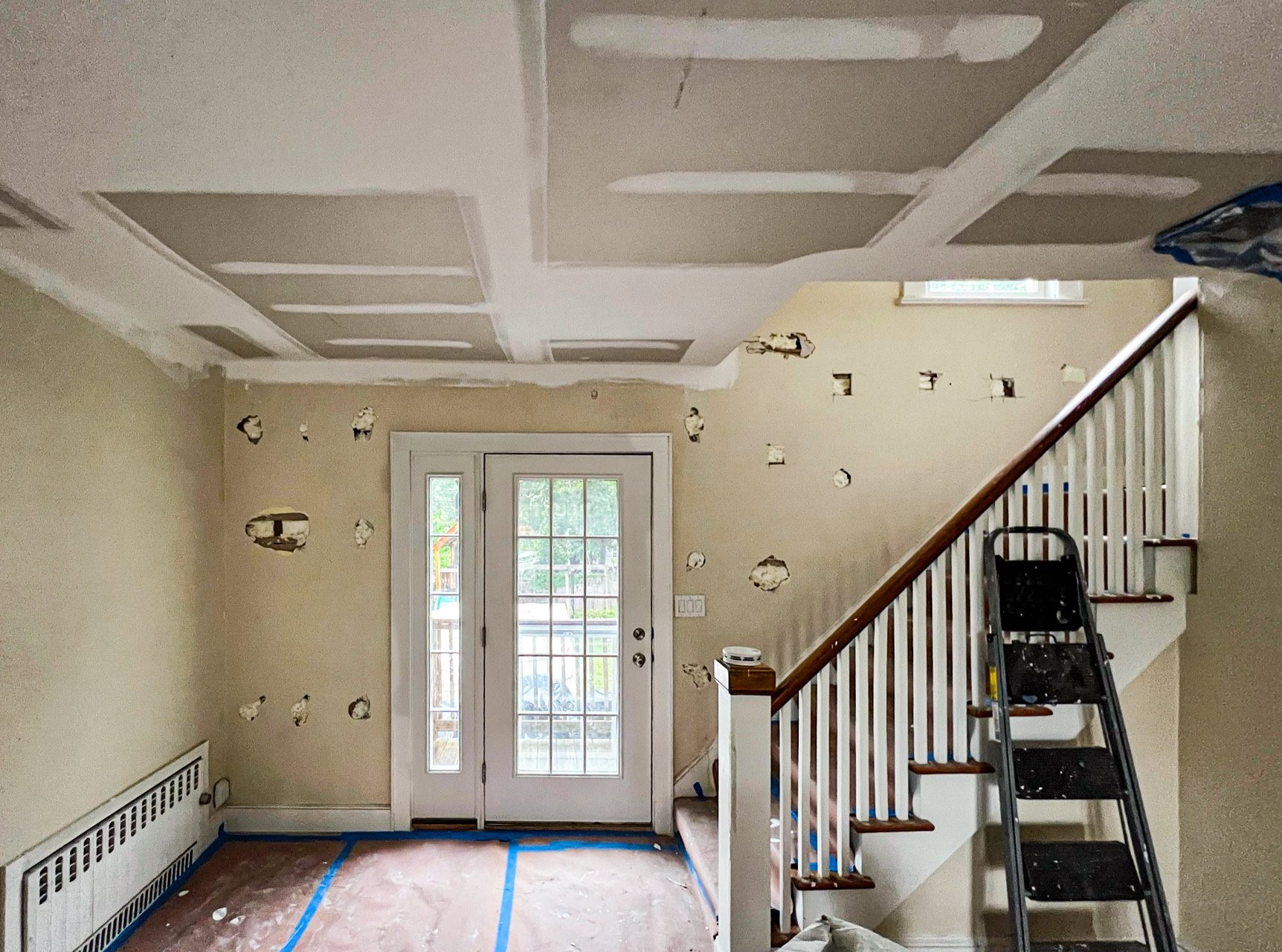 Interior of a house under renovation with drywall installation, a ladder, a door leading outside, and holes in the wall.