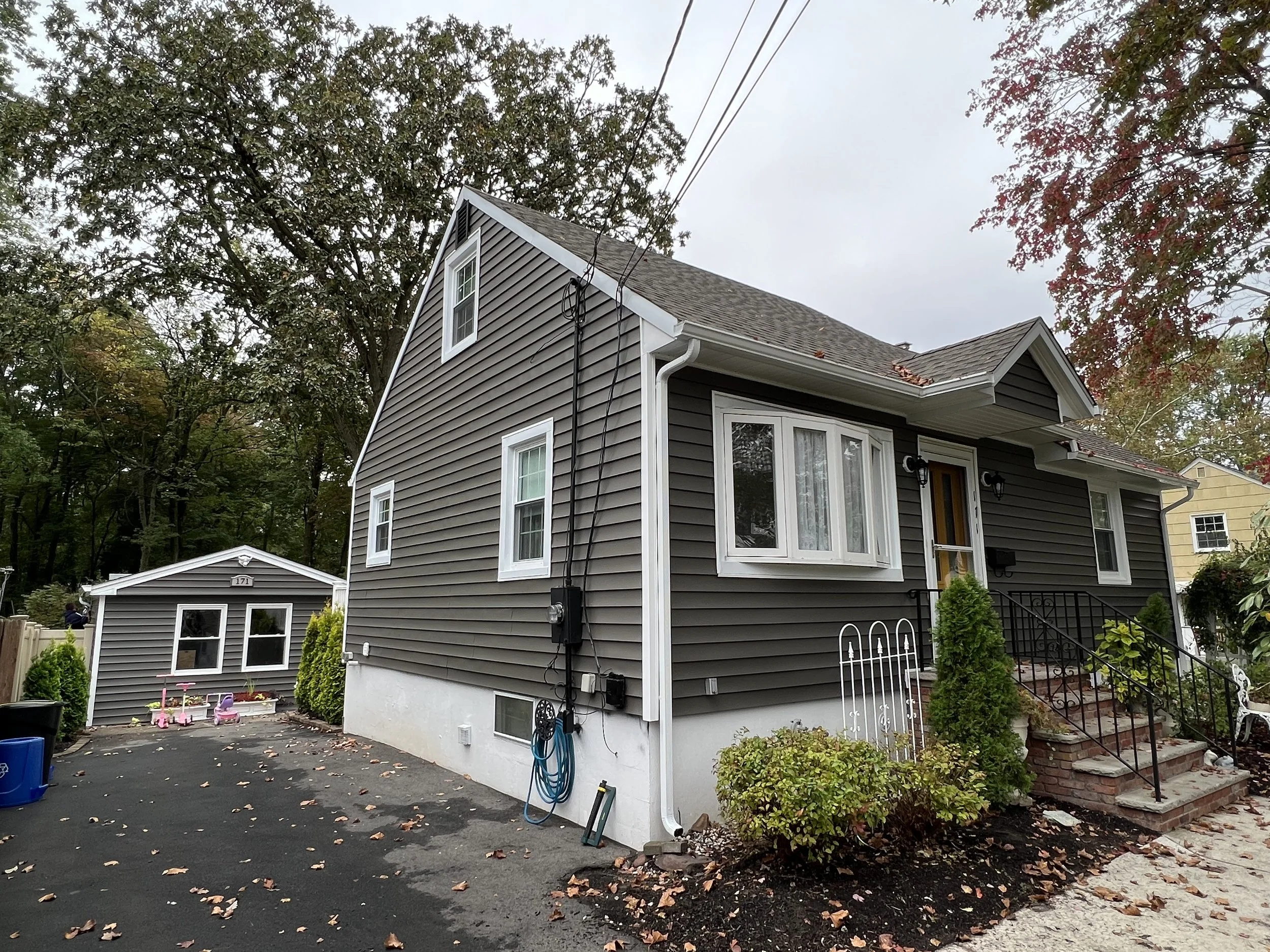 A dark gray house with white trim and a small front porch with stairs, surrounded by trees with autumn leaves and a driveway with fallen leaves.