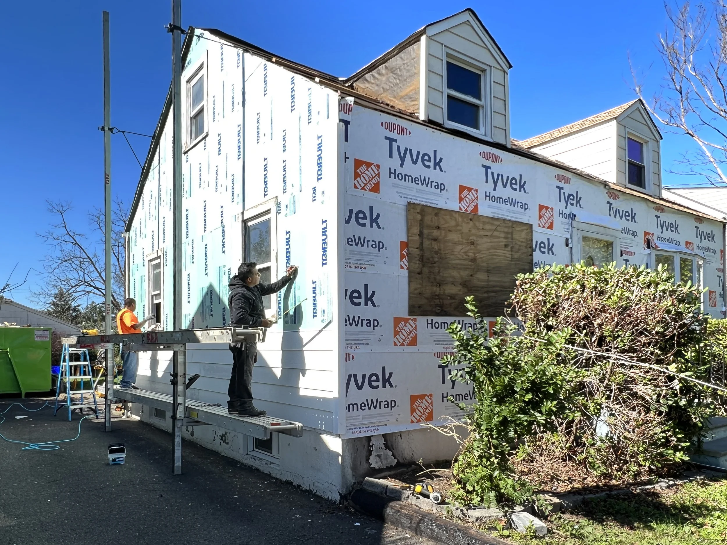 Construction workers installing siding on a house with Tyvek HomeWrap, a large bush in the foreground, and a clear blue sky.