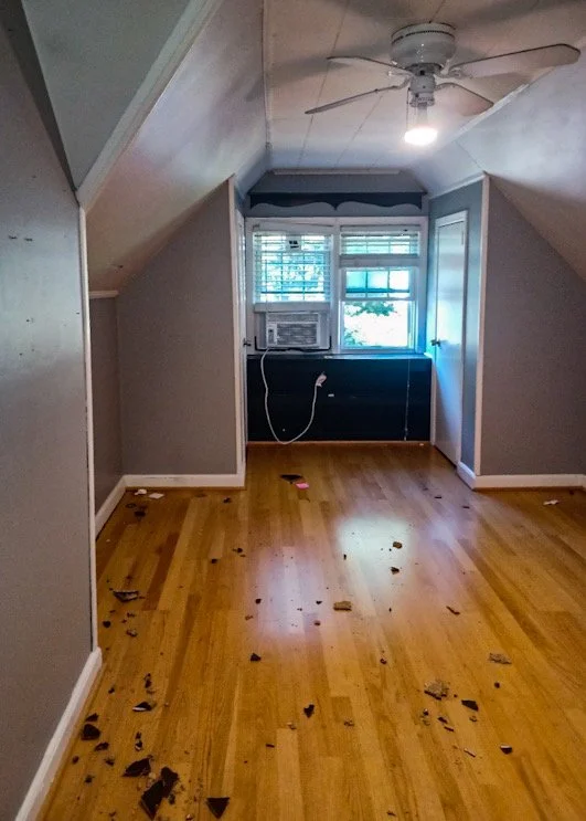 Empty room with wooden floor and gray walls, scattered pieces of debris on the floor, a ceiling fan, and an air conditioning unit installed in a front window.