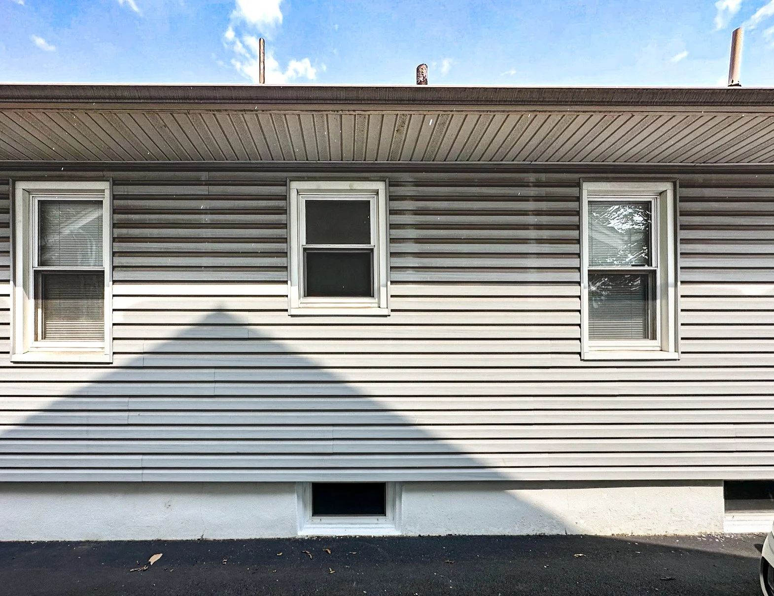 Front exterior of a house with beige horizontal siding, three windows (two on upper level and one basement vent), brown roof, and a shadow cast on the siding. The sky is blue with some clouds.