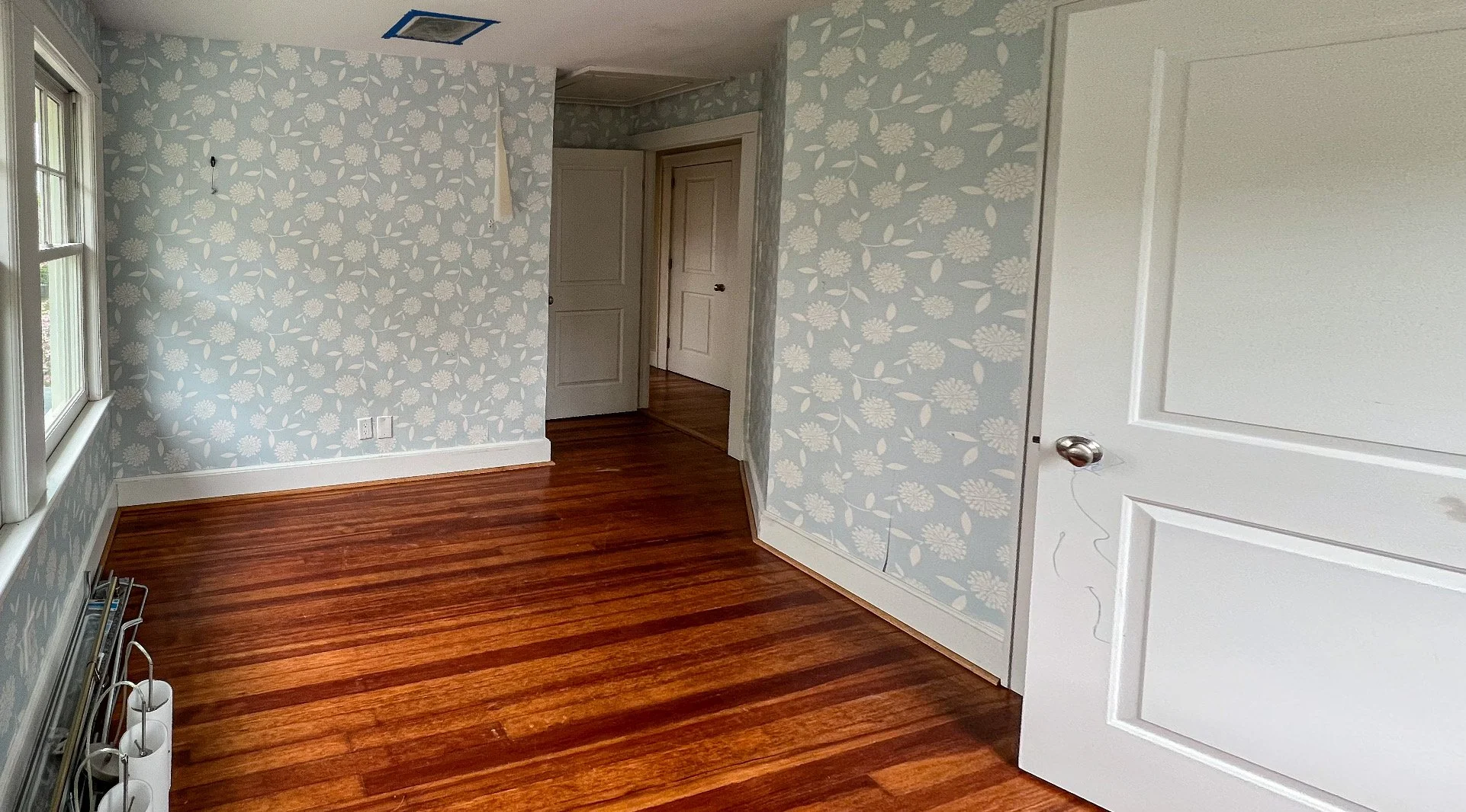 Unfurnished room with hardwood floors, blue floral wallpaper, a window, a white door, and a small section of a white wall.