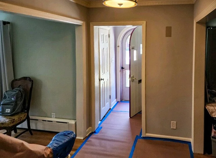 Interior of a house near the front door, with blue painter's tape on the floor and door frame, a chair, a backpack, and a baseboard heater.