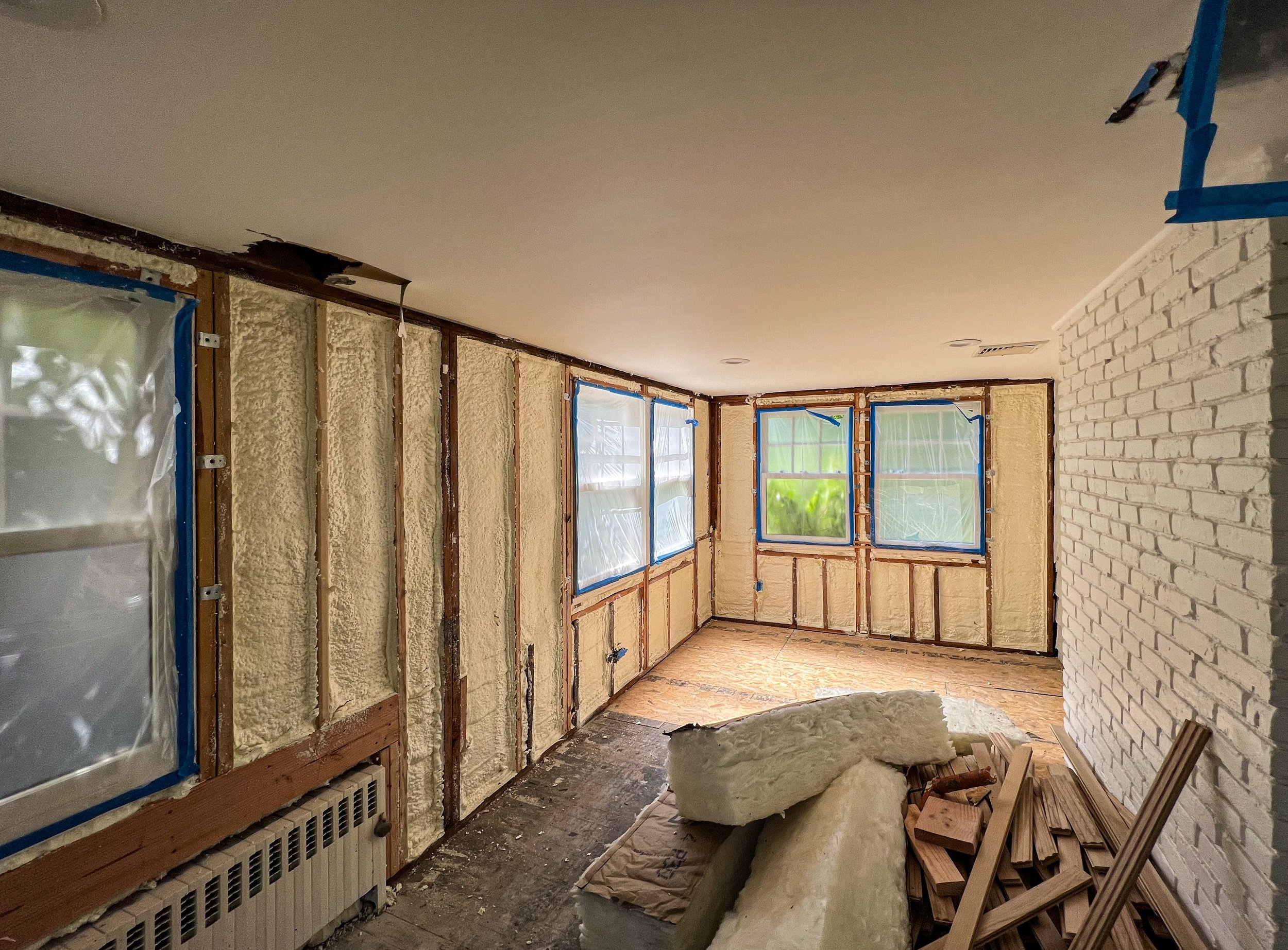 Room under renovation with insulation, windows covered with protective plastic, and construction materials like foam and wood on the floor.