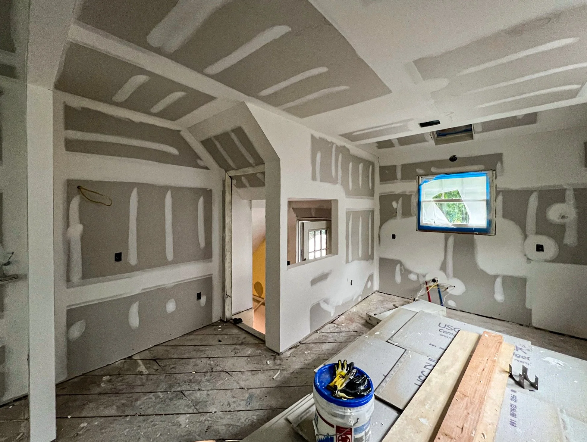 Room under construction with drywall installed, taped, and mudded. Construction tools and materials on the floor, including a bucket, gloves, and wooden planks. Window with protective covering.