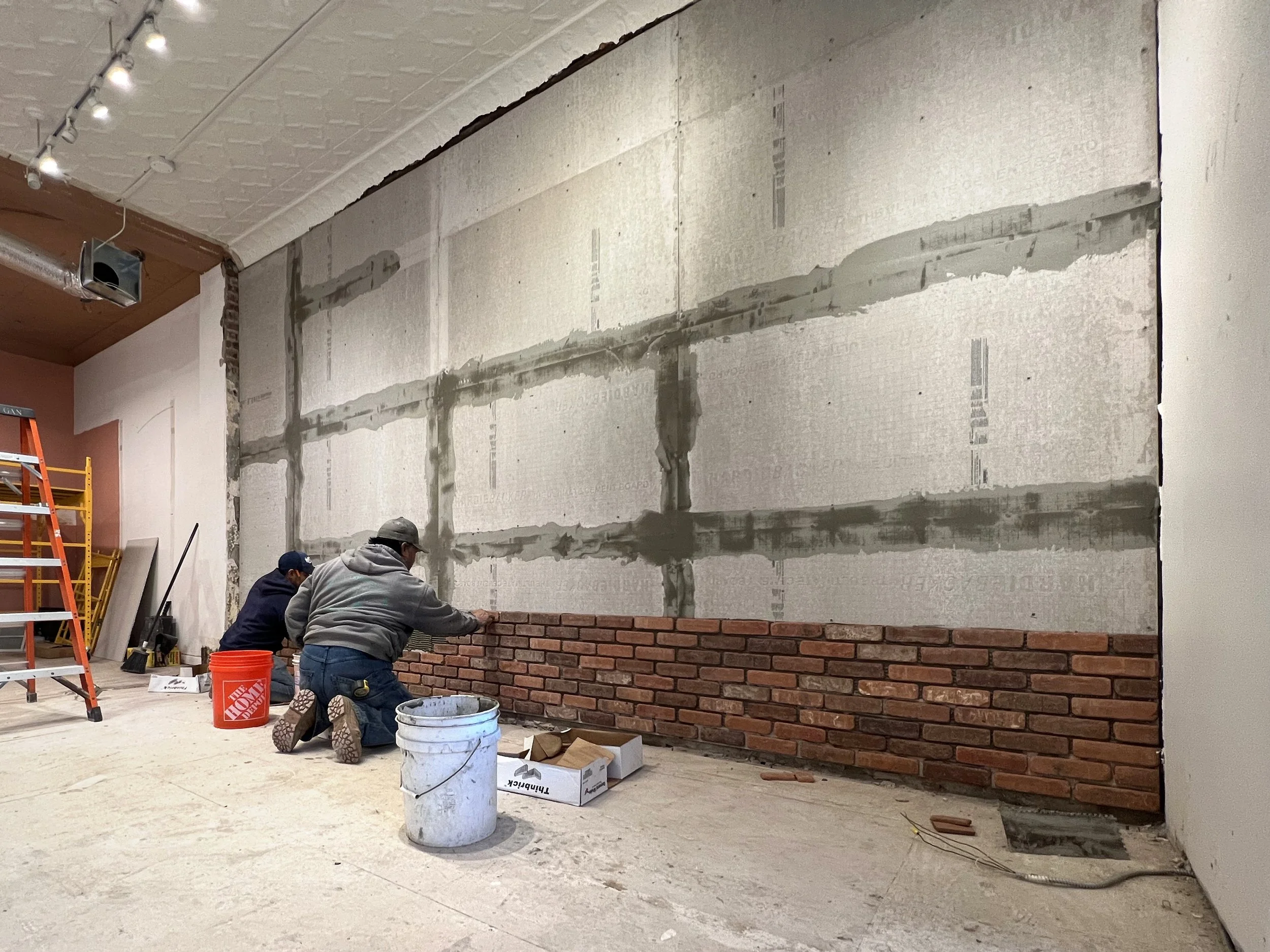 Two construction workers kneeling and laying bricks along a wall inside a building under renovation. The wall background shows insulation and unfinished sections.