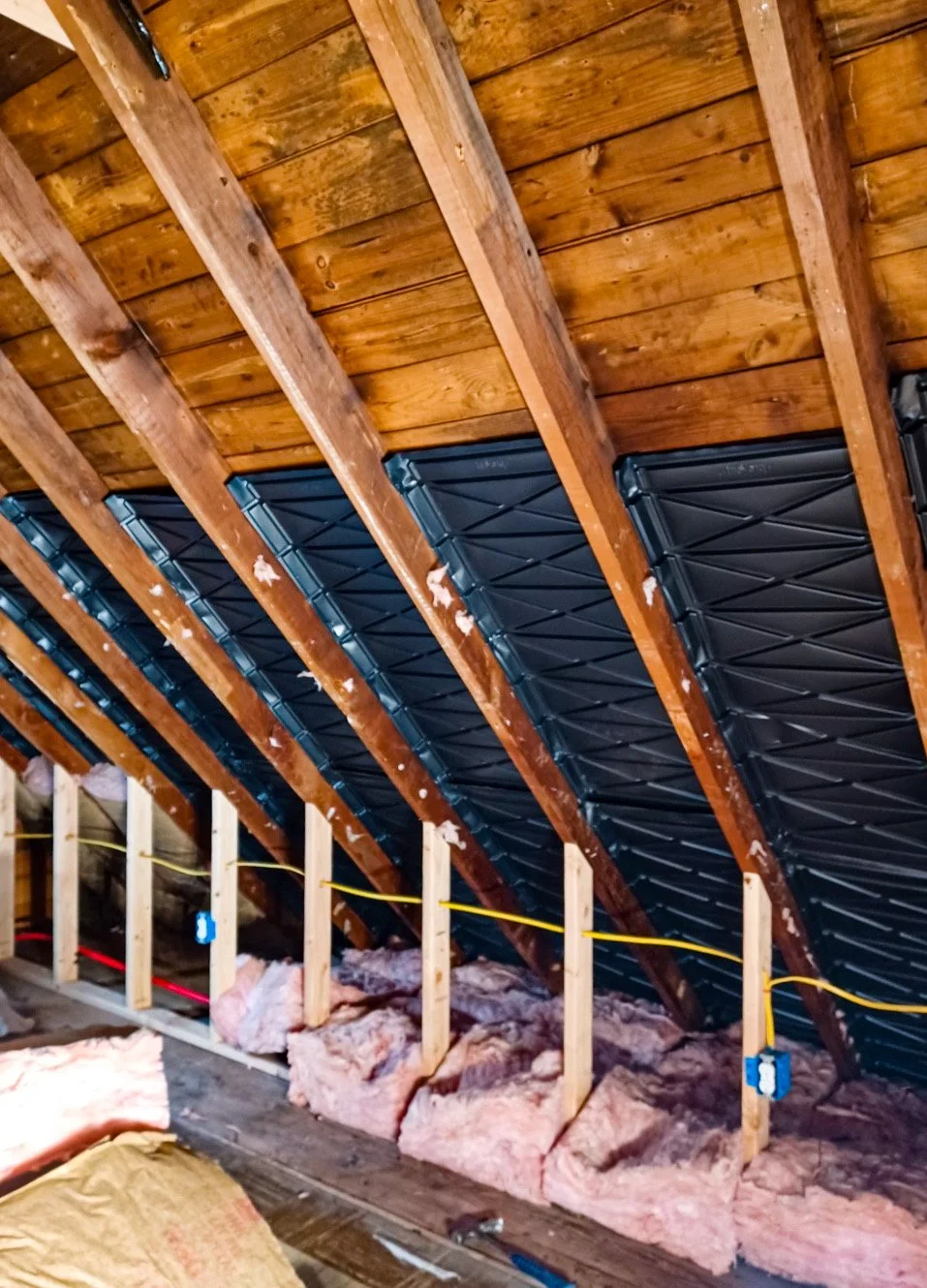 Insulation being installed in an attic with exposed wooden rafters, blue waterproof membrane, and electrical wiring.