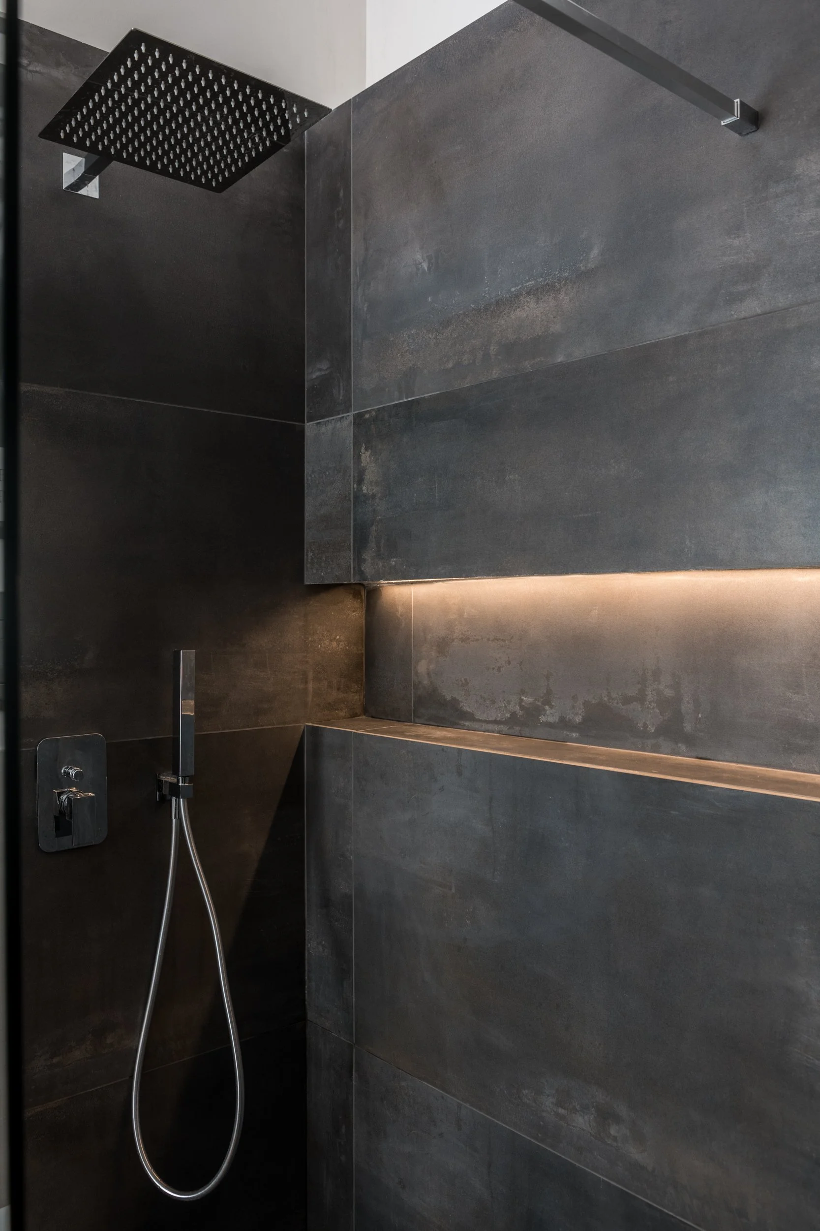 Doccia walk-in con parete in vetro, rivestimento scuro effetto metallo e illuminazione LED integrata in un bagno contemporaneo.