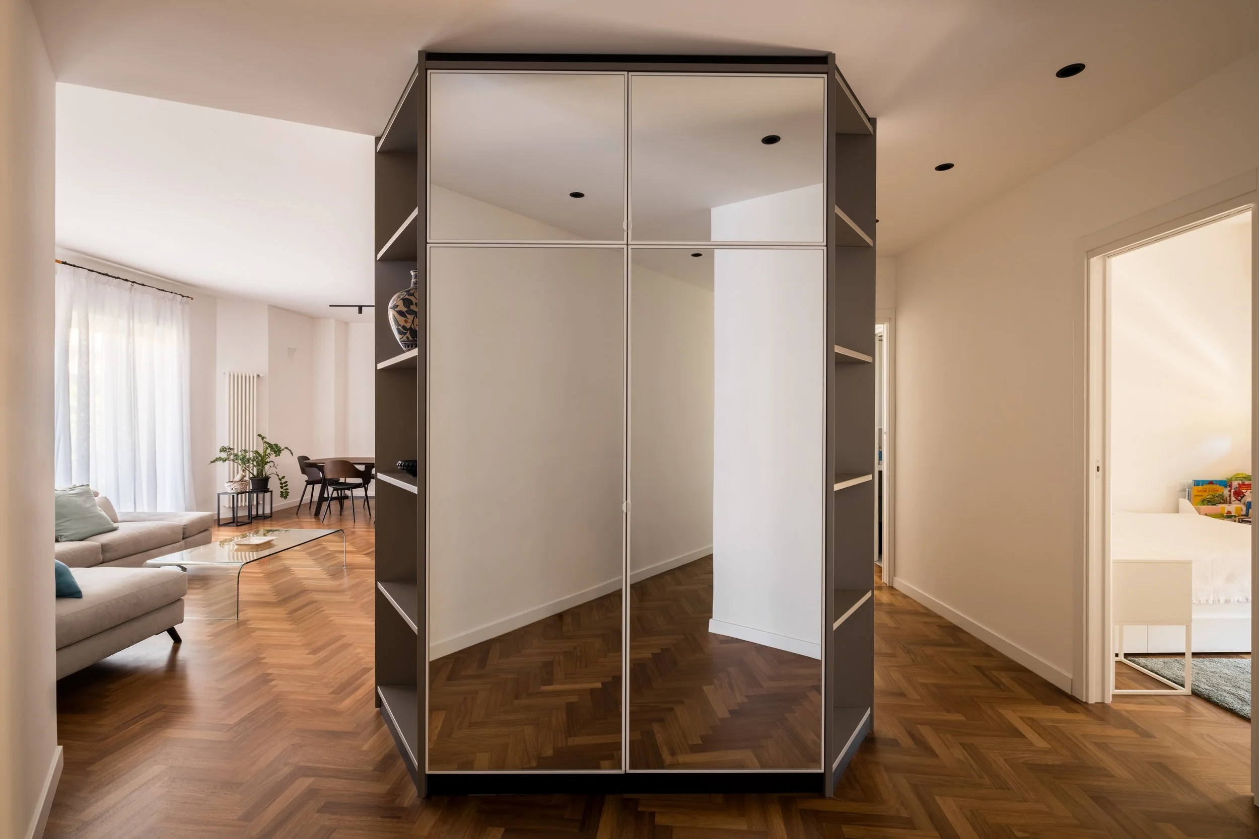 Le ante specchiate del volume su misura trasformano il disimpegno in uno spazio dinamico e percettivamente ampliato, moltiplicando le prospettive e riflettendo la continuità del parquet in iroko. Le superfici riflettenti attenuano la presenza del vol