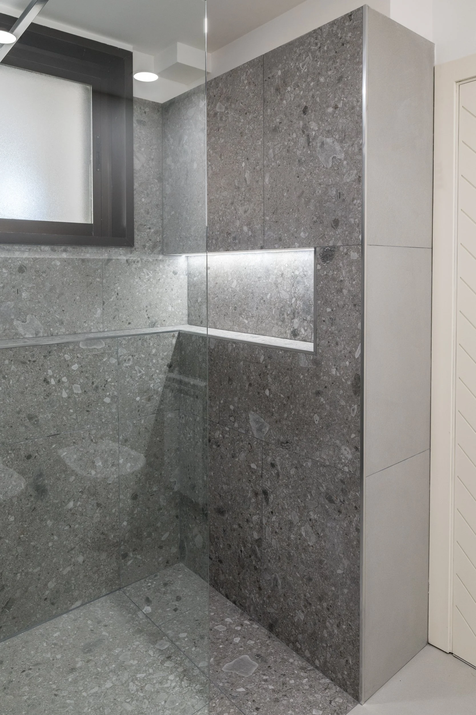 Doccia walk-in con rivestimento in gres materico e nicchia illuminata nella villa a Palermo. La doccia walk-in è progettata come uno spazio continuo rivestito in gres materico. La nicchia illuminata e la lastra in vetro contribuiscono a una percezion
