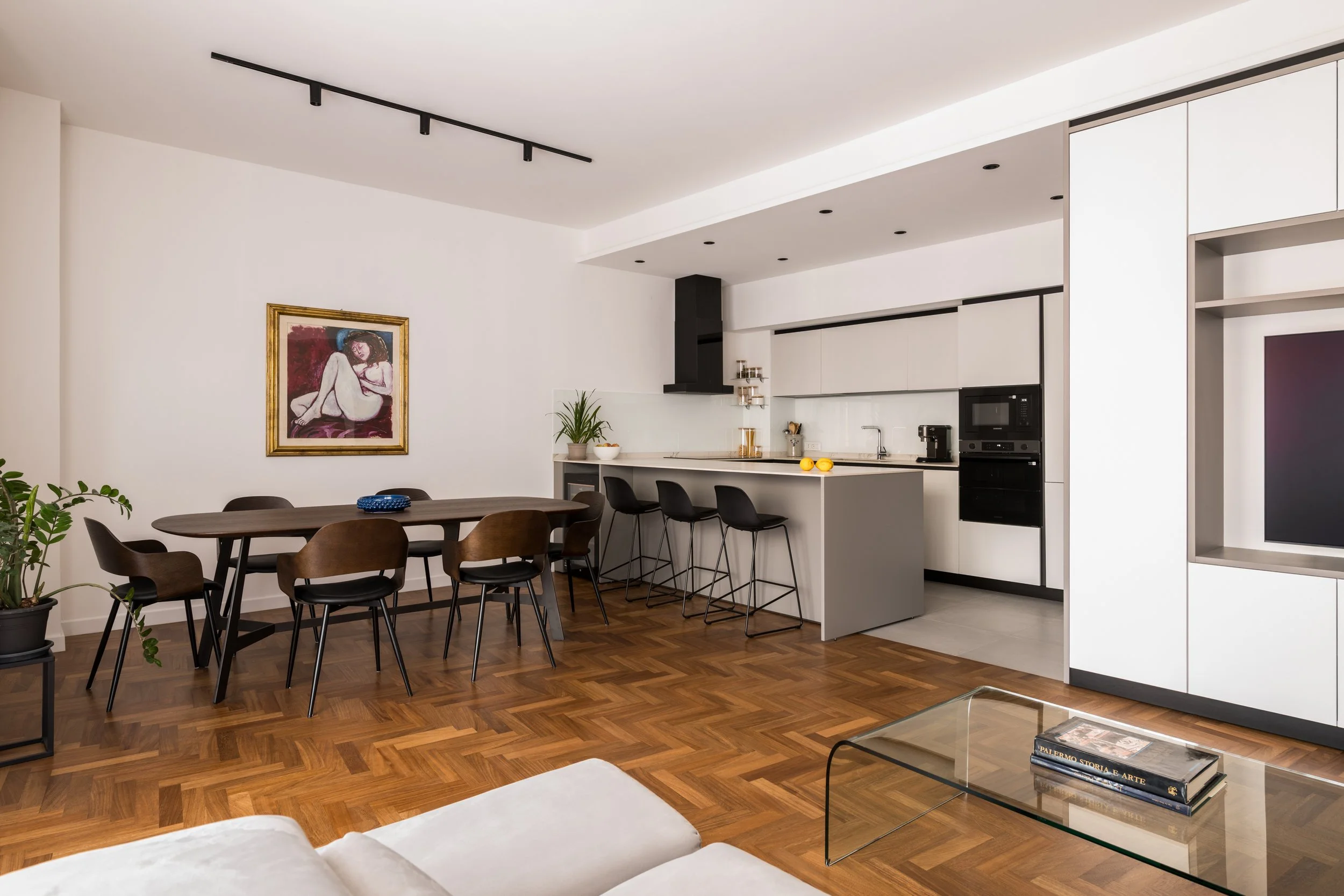 La zona giorno si sviluppa come un unico ambiente fluido in cui cucina, sala da pranzo e soggiorno dialogano attraverso una composizione rigorosa di volumi, materiali e proporzioni. La cucina Elmar con isola si integra nel sistema di arredi su misura