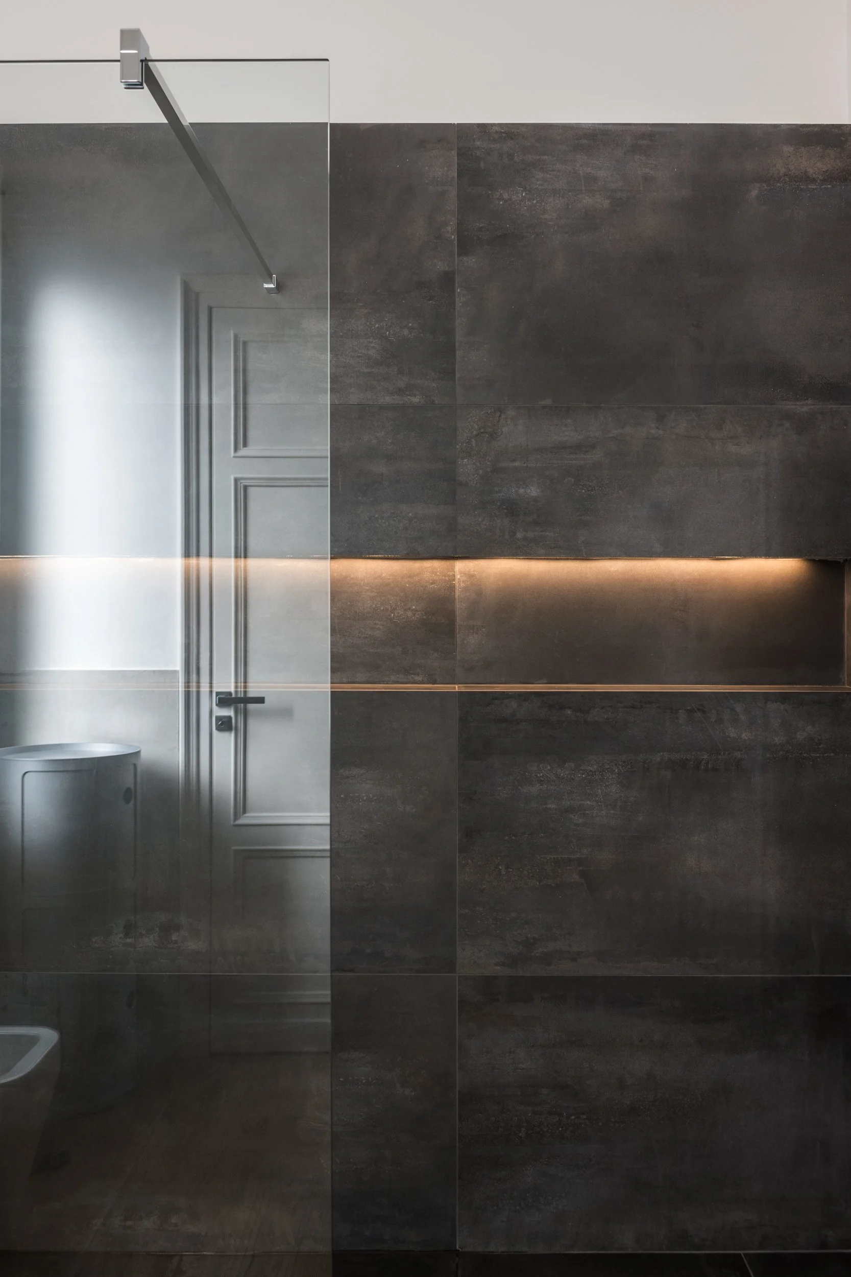 Bagno contemporaneo dell'appartamento Verticalismi a Palermo. Rivestimenti materici scuri, superfici continue e illuminazione integrata definiscono uno spazio essenziale in cui luce e materia costruiscono profondità e atmosfera.