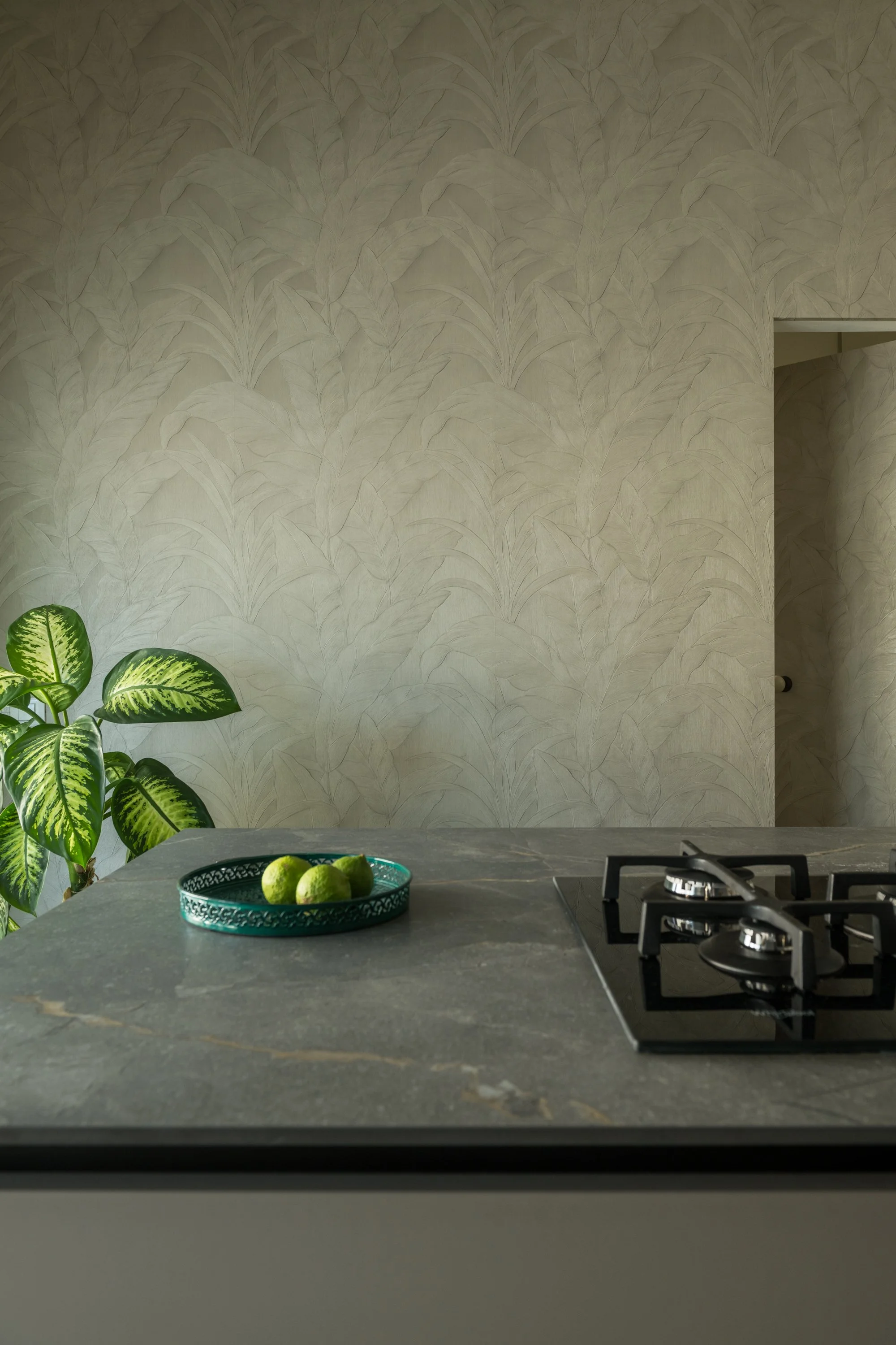 Dettaglio di una cucina contemporanea a Mondello, Palermo, con parete rivestita da carta da parati botanica e porta filomuro integrata nella decorazione. Il piano cucina in finitura effetto pietra e la vegetazione contribuiscono a una composizione ma