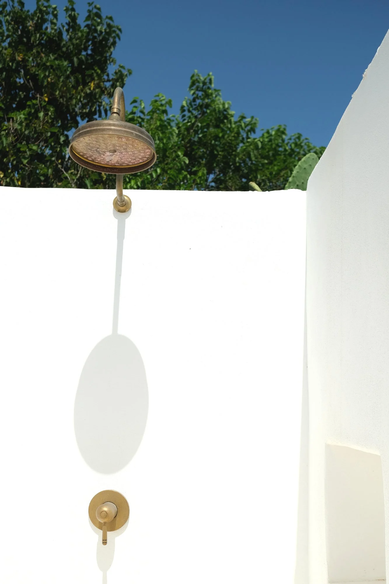 Doccia esterna con soffione in ottone su pareti bianche intonacate illuminate dal sole mediterraneo.