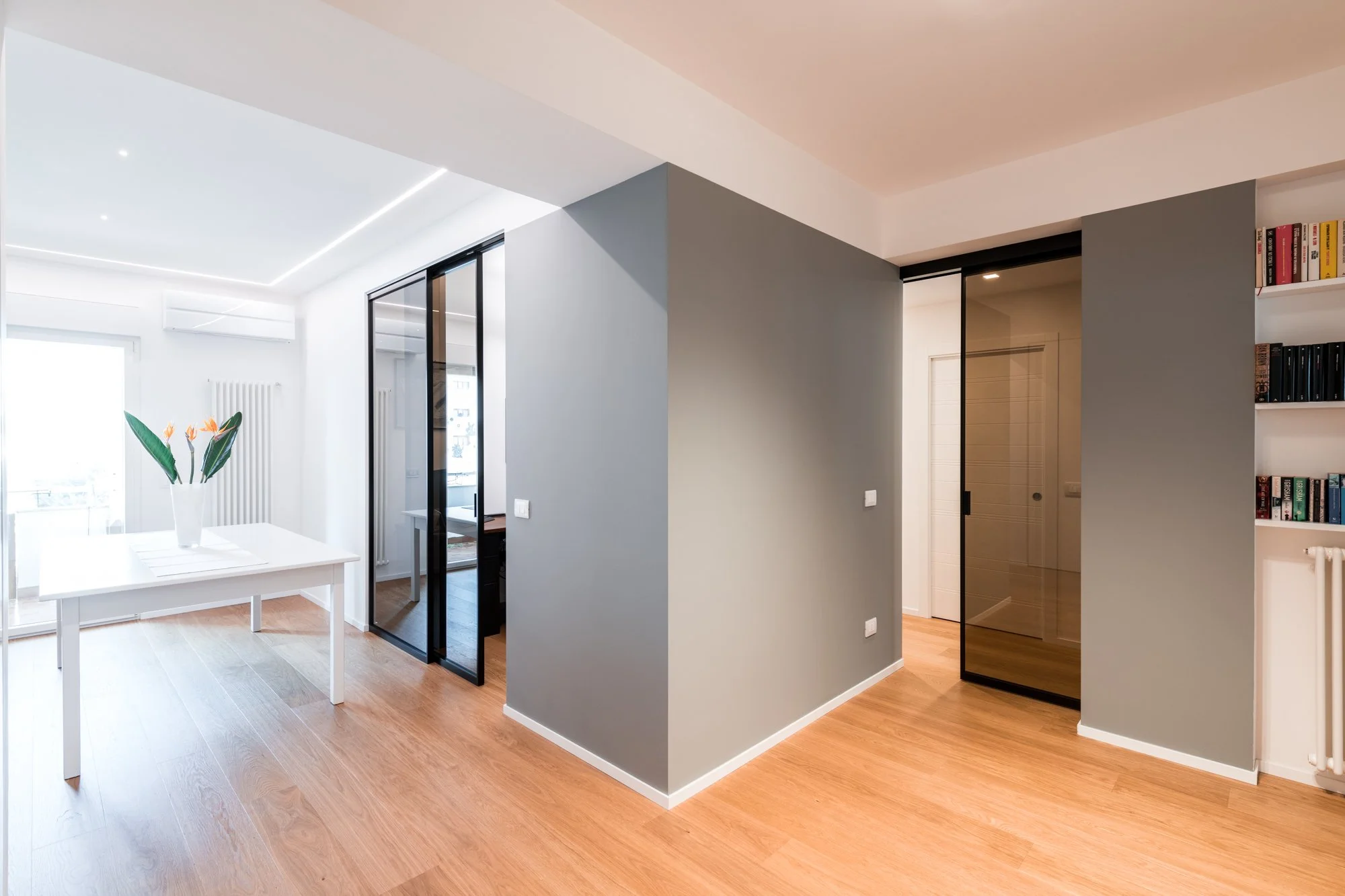 Vista dell’ingresso dell’appartamento Mikado a Palermo. Il volume grigio centrale organizza i percorsi tra zona giorno, studio e zona notte, mentre parquet in rovere, porte vetrate fumé e illuminazione integrata definiscono le gerarchie dello spazio.