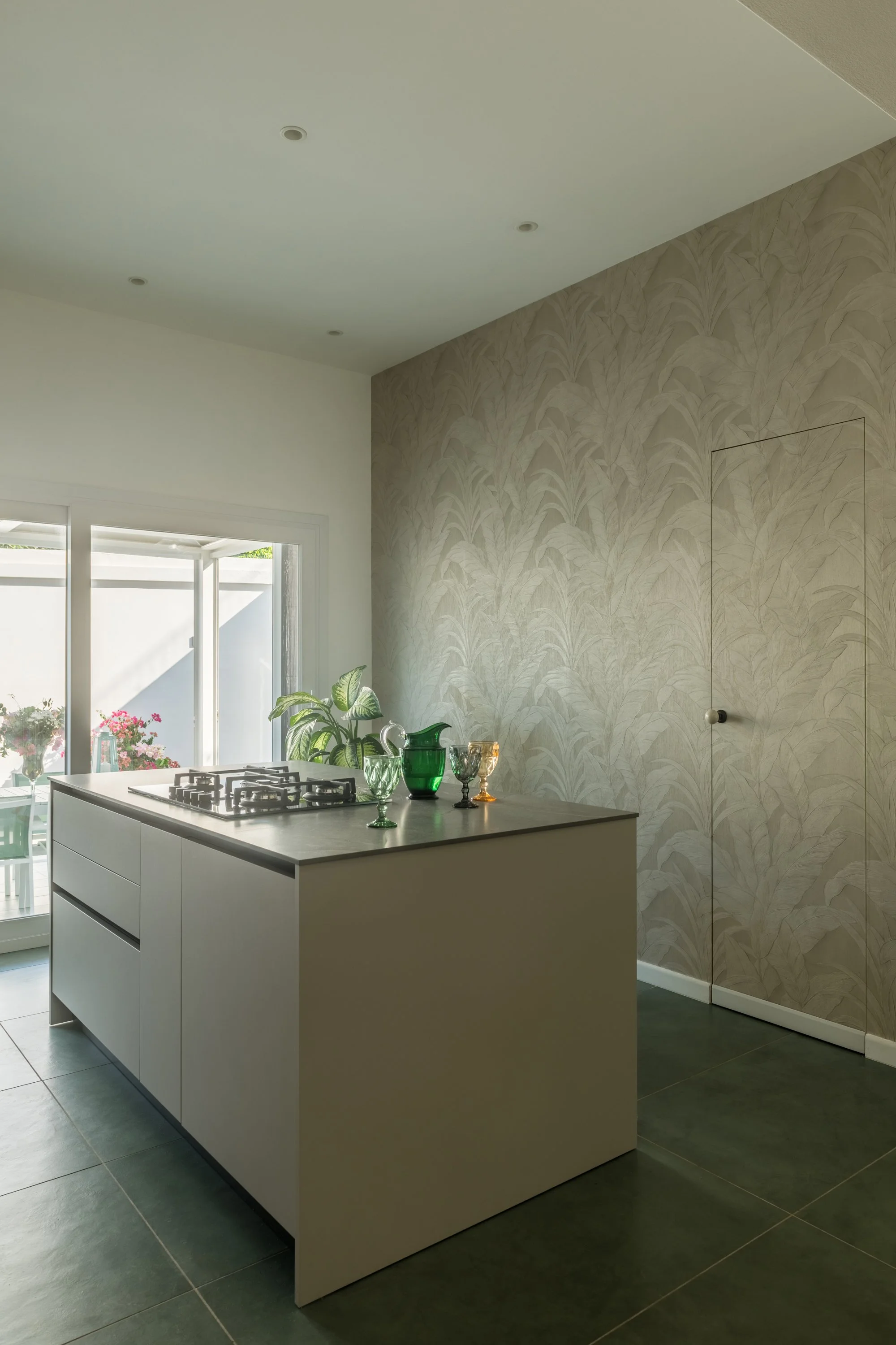 Cucina contemporanea a Mondello, Palermo, caratterizzata da un'isola centrale minimalista, piano effetto pietra e parete decorata con carta da parati a motivi floreali e botanica. La composizione integra una porta filomuro rivestita con lo stesso mot
