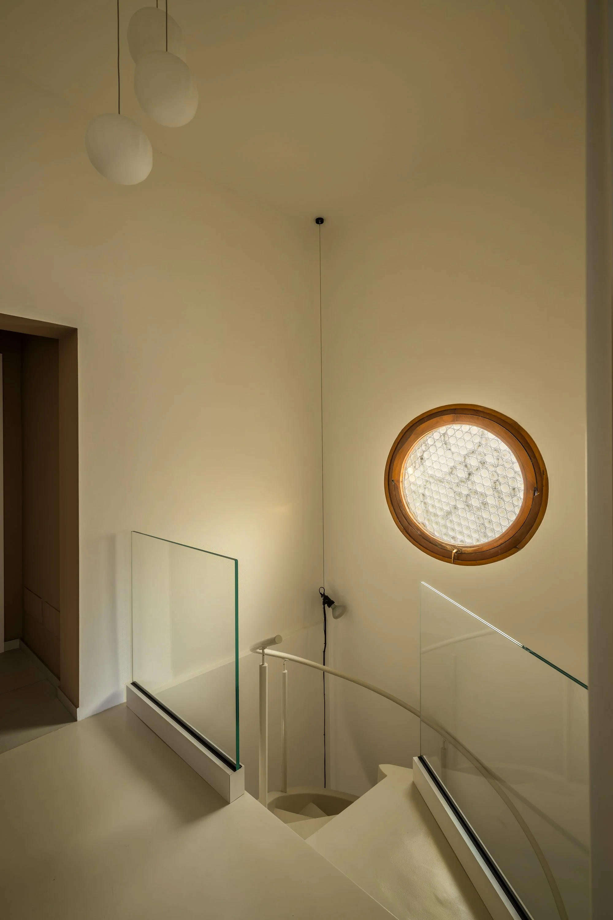Vista dal pianerottolo della zona notte di una scala elicoidale interna resinata color avorio con parapetti in vetro, corrimano curvo e oblò circolare in legno sulla parete. Le superfici color crema, le lampade sospese e la luce naturale costruiscono