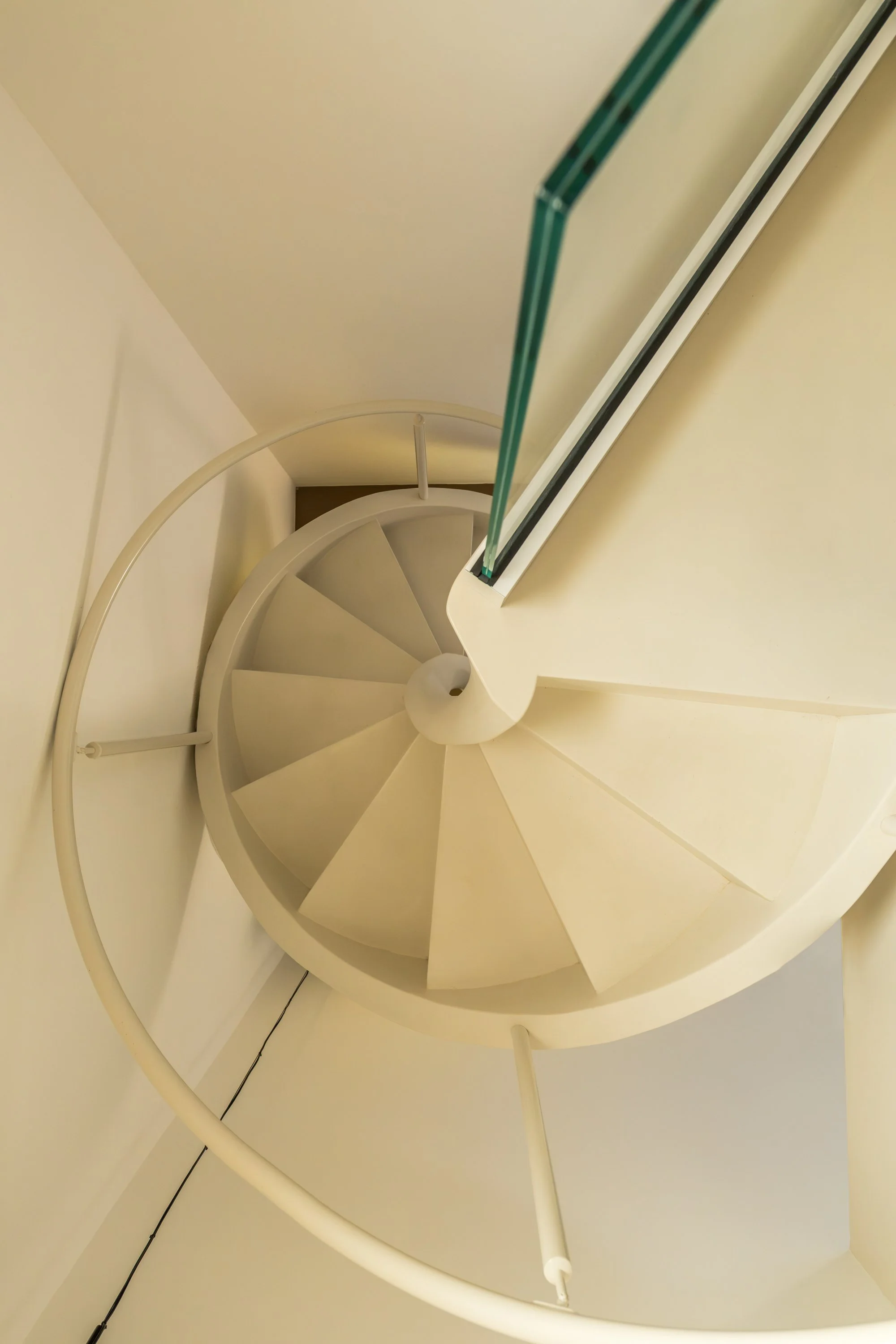 Dettaglio di una scala elicoidale resinata color avorio con parapetto in vetro trasparente e corrimano curvo, inserita in un interno dalle tonalità calde e minimali. La geometria circolare della scala definisce lo spazio verticale e crea un forte ele