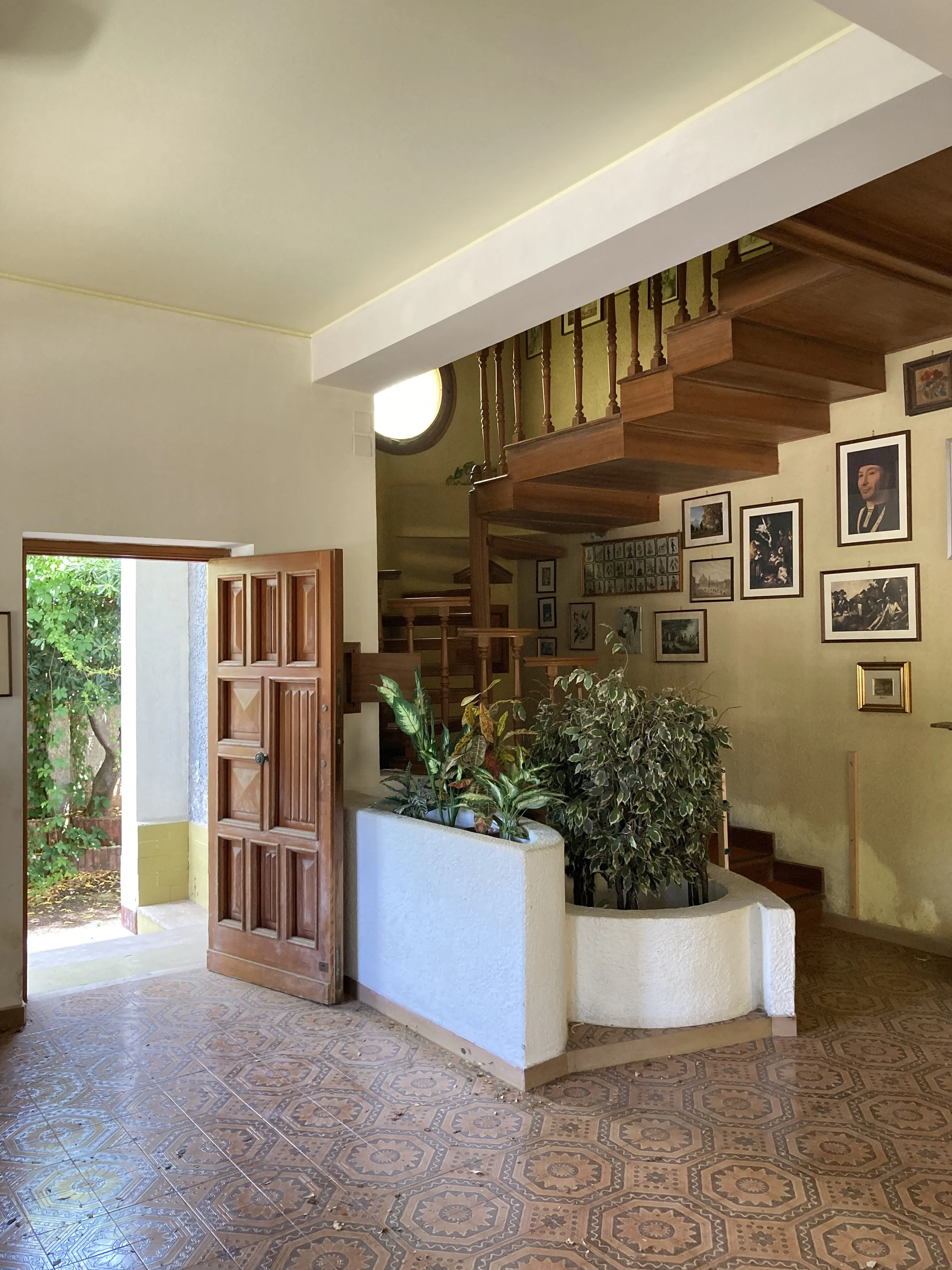 Vista dell'ingresso della villa e della scala originaria in legno prima della trasformazione dell'edificio. Il progetto sostituisce il collegamento verticale esistente con una nuova scala elicoidale rivestita in resina, posta nel baricentro della pia