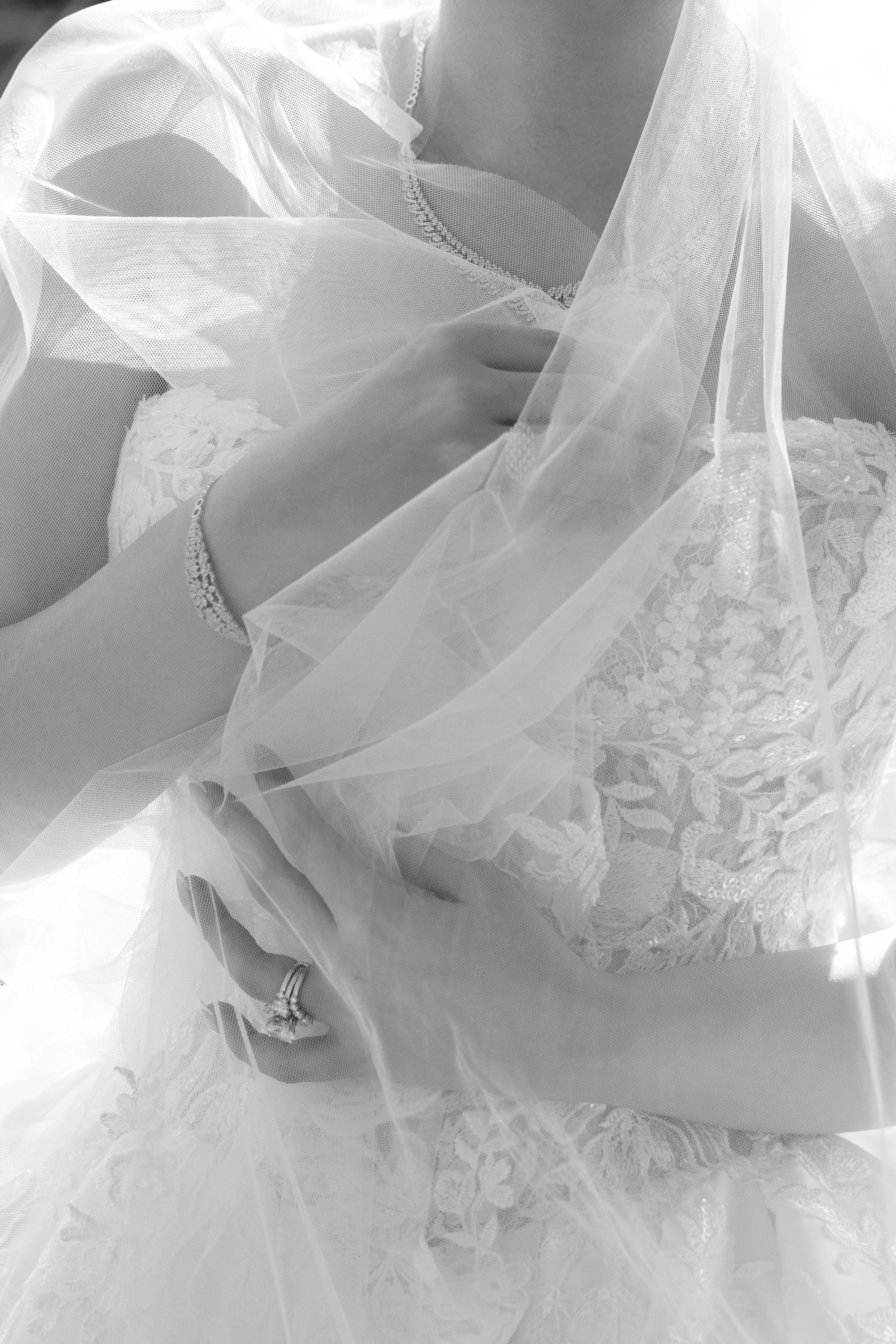 Close-up of a woman in a wedding dress with lace details, holding her veil and showing a wedding ring on her finger.