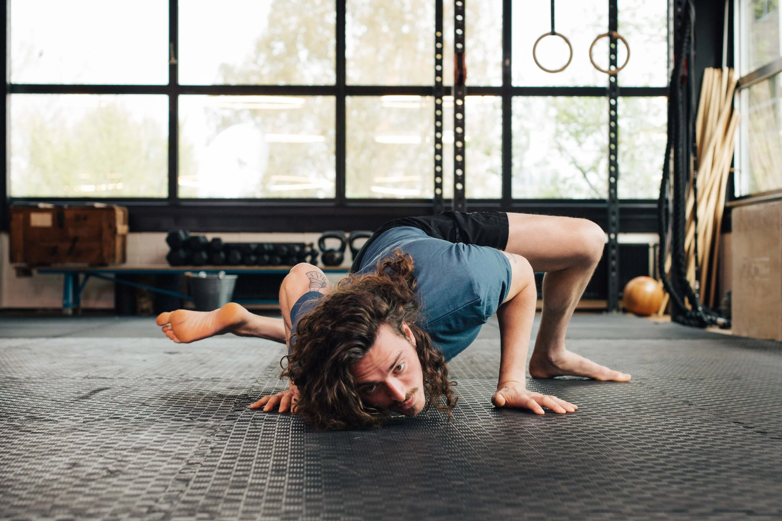 Man with curly hair in a gym performing an exercise on the floor, with gym equipment and windows in the background.