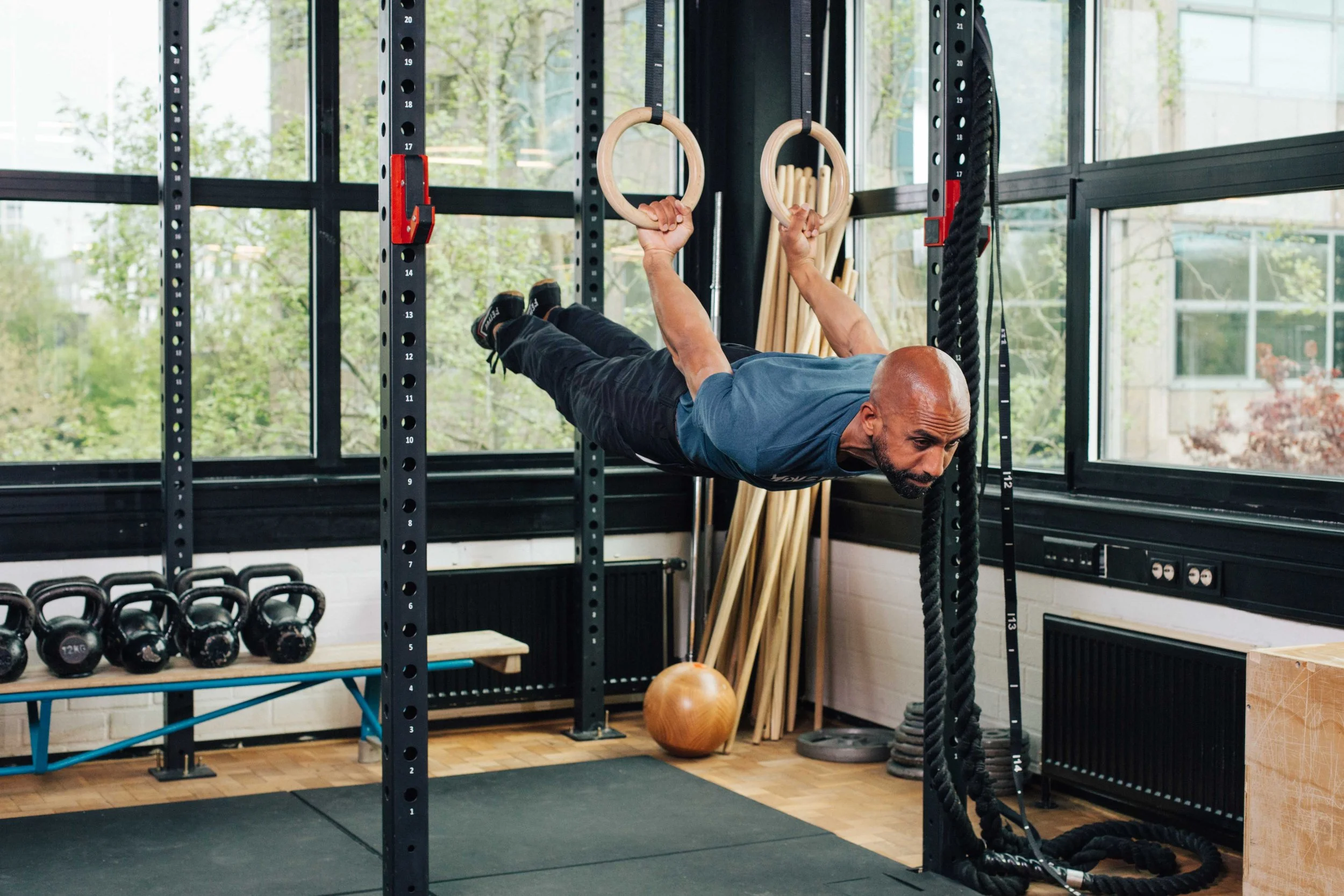 Man performing a horizontal hold using gymnastic rings at a gym with kettlebells, wooden sticks, and exercise equipment in the background.