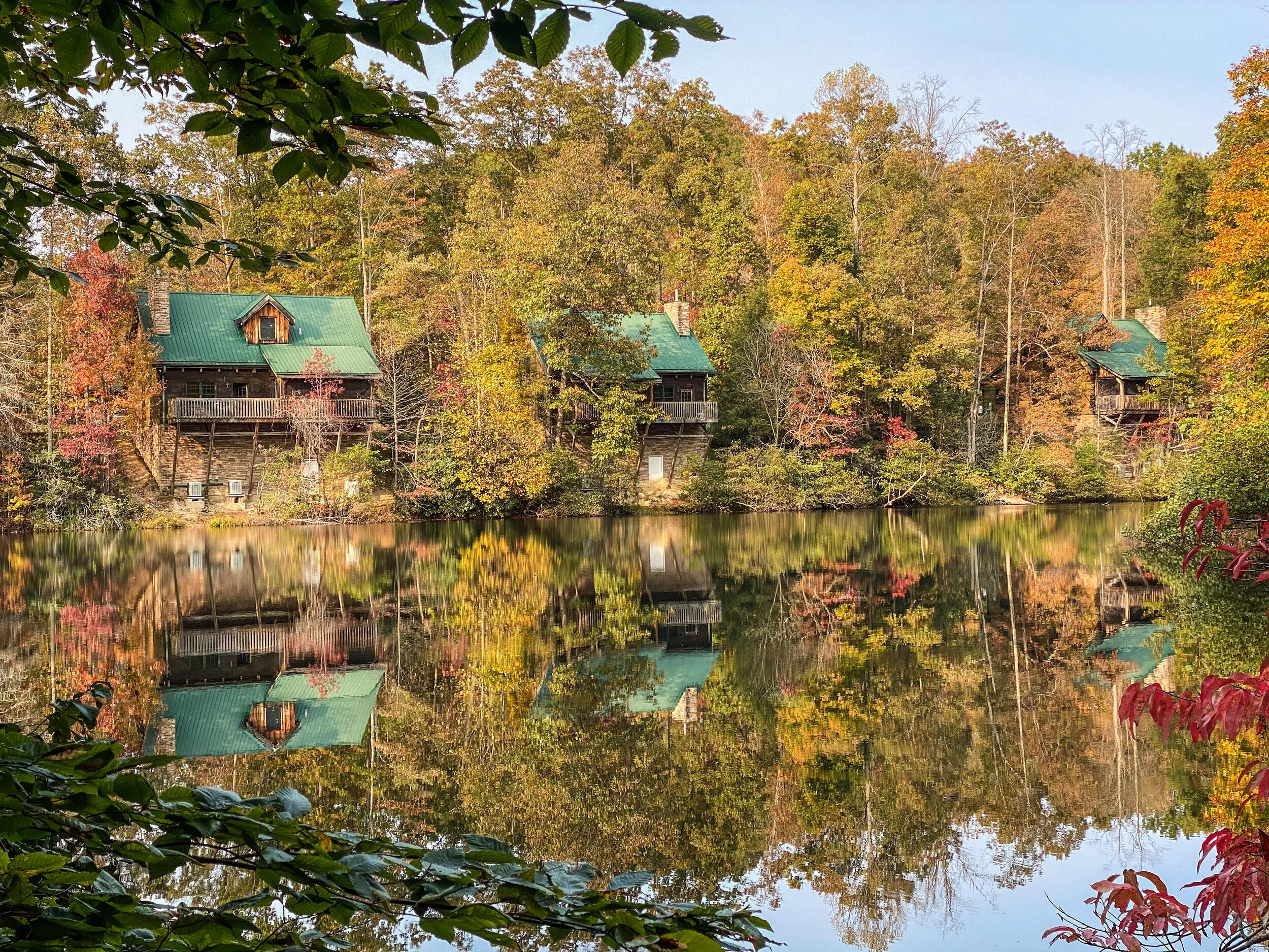Three cabins with green roofs on a hillside by a lake, reflecting the autumn-colored trees and cabins in the calm water.