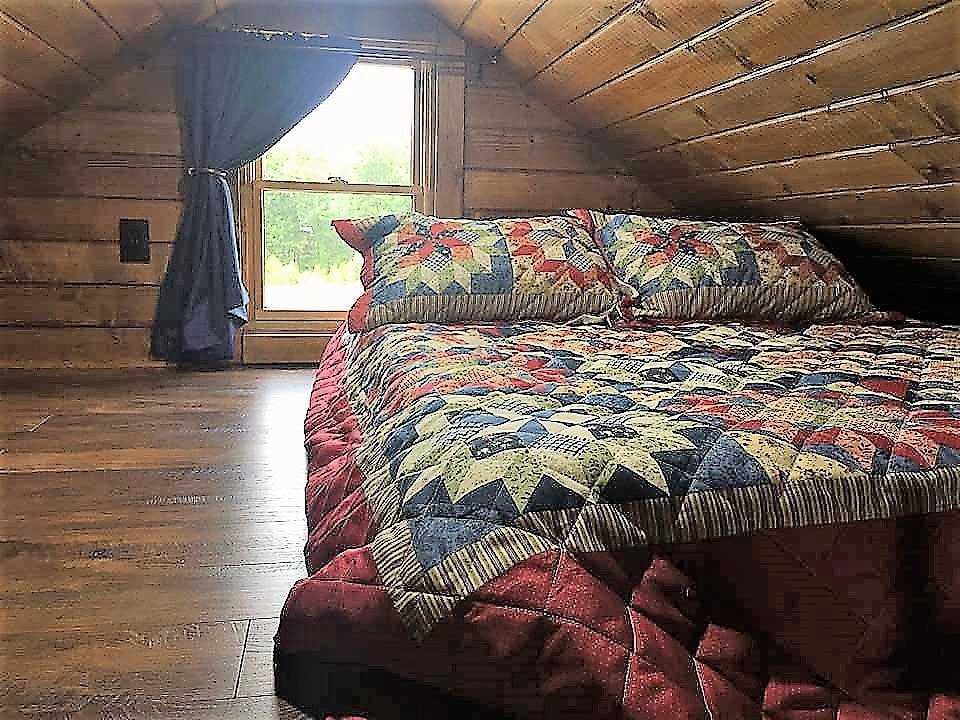 A cozy rustic bedroom with wooden walls and ceiling, a bed with quilted bedding, and a window with blue curtains.