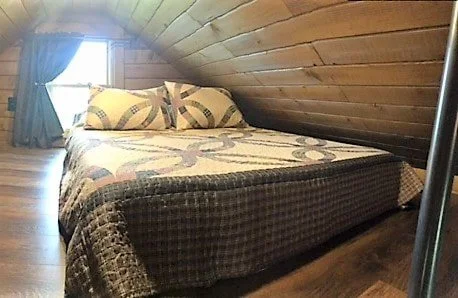 A bed with a quilt and pillows in a cozy attic room with wooden ceiling and a small window with curtains.