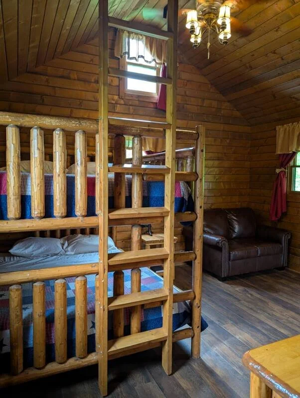 Interior of a rustic wooden cabin bedroom with a set of bunk beds made of logs, a leather sofa, a small window with curtains, and a wooden ceiling with a chandelier.