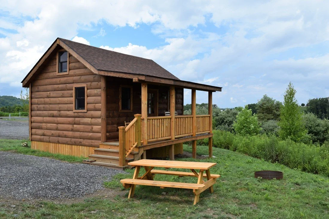 A small wooden cabin with a porch and a picnic table outside, surrounded by greenery and a partly cloudy sky.