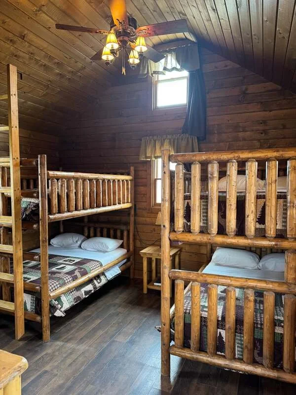 A rustic wooden cabin bedroom with two sets of bunk beds, a small nightstand, wooden walls, and a ceiling fan with lights.
