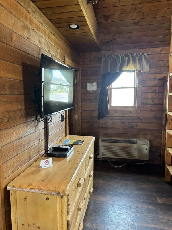 Wooden interior room with a mounted flat-screen TV, a wooden dresser with a remote and pamphlets, a window with curtains, and a wall-mounted air conditioner.