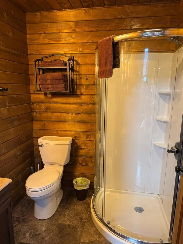 Bathroom with wood-paneled walls, a white toilet, a wall-mounted shelf holding folded towels, a small trash can, and a glass-enclosed shower stall.