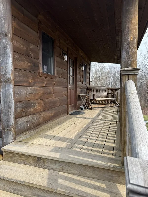 Wooden porch with bench swing, small table, and steps leading to the yard, attached to a log cabin-style house.