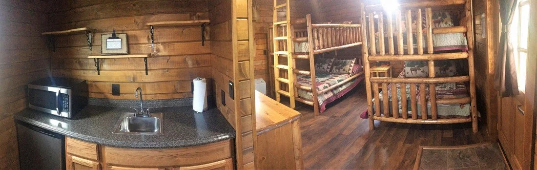 Interior of a rustic cabin with a small kitchen area featuring a microwave, sink, and countertop, and a sleeping area with wooden bunk beds and a small seating area. Wood paneling is throughout the space.