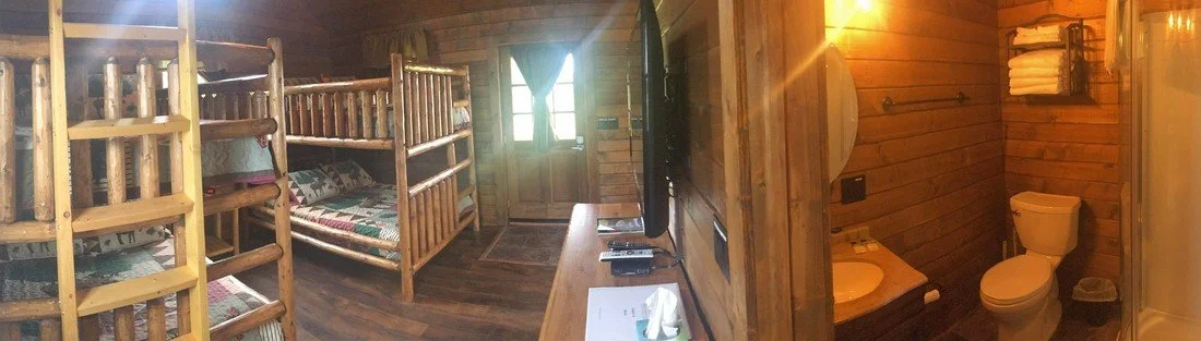 A rustic cabin bedroom with wooden bunk beds on the left, a door at the far end, a small television and a dresser on the right, and an adjacent bathroom with a toilet and sink.