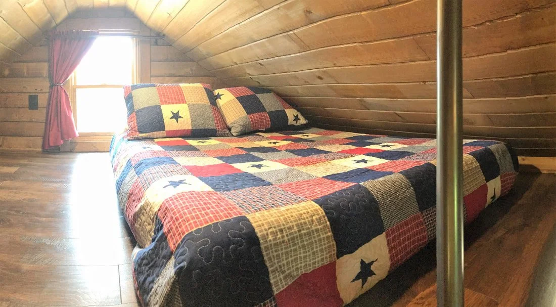 A cozy bedroom with a sloped wooden ceiling, a window with pink curtains, and a bed with a colorful quilt and matching pillows.