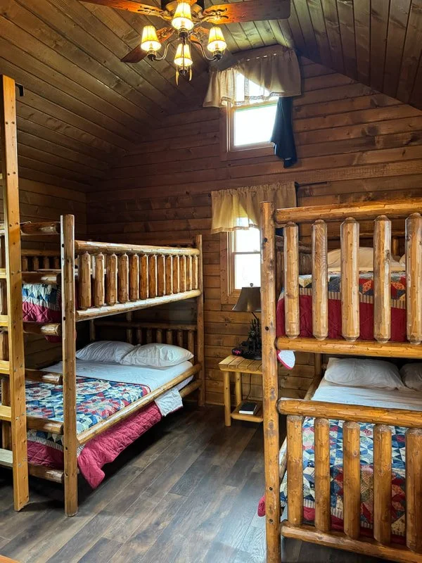 A cozy room with wood-paneled walls and ceiling, featuring two sets of wooden bunk beds with quilted mattresses, a small nightstand with a lamp, and small windows with curtains, illuminated by a ceiling fan with lights.
