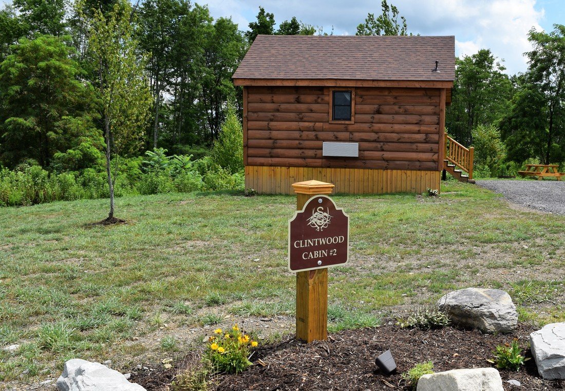 A small wooden cabin with a brown shingled roof and a small window, surrounded by green trees and grass. In front of the cabin, there is a sign that reads "Clintwood Cabin #2," and small flower beds with yellow flowers and rocks.
