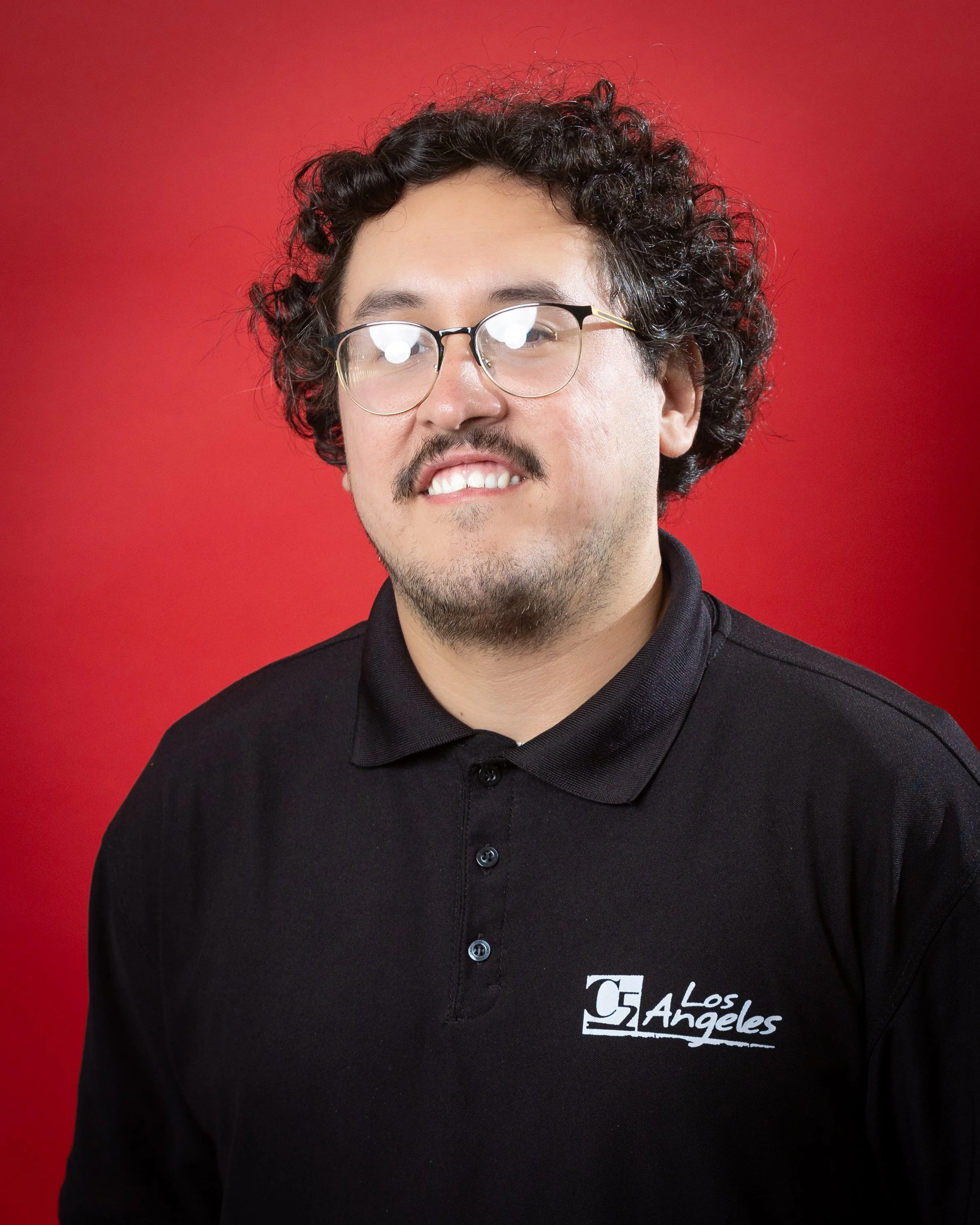 A young man with curly hair and glasses wearing a black polo shirt with the C5 Los Angeles logo.