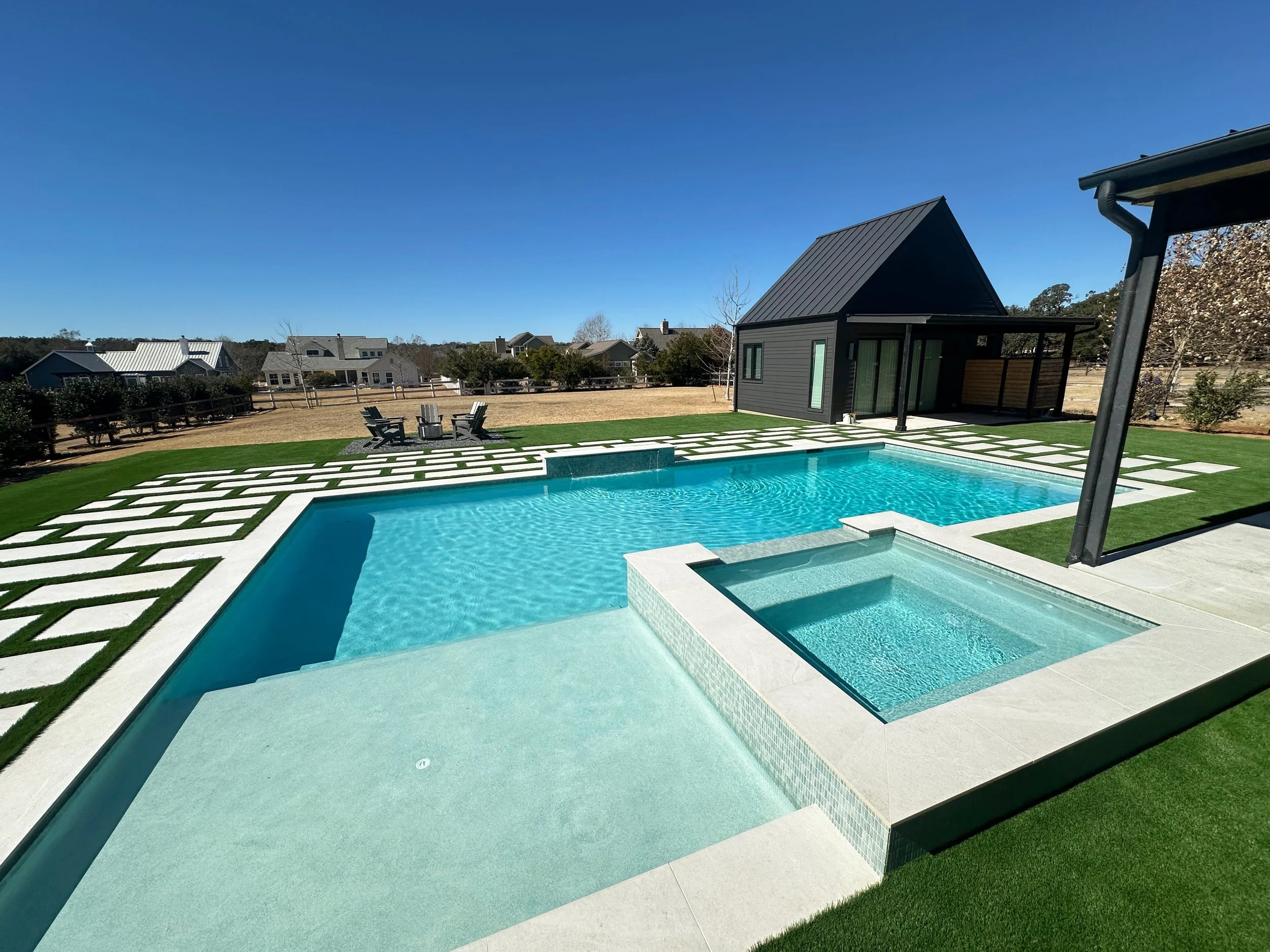 Modern farmhouse pool with tanning ledge, sun shelf, and raised spillover spa beside a black metal-roof casita