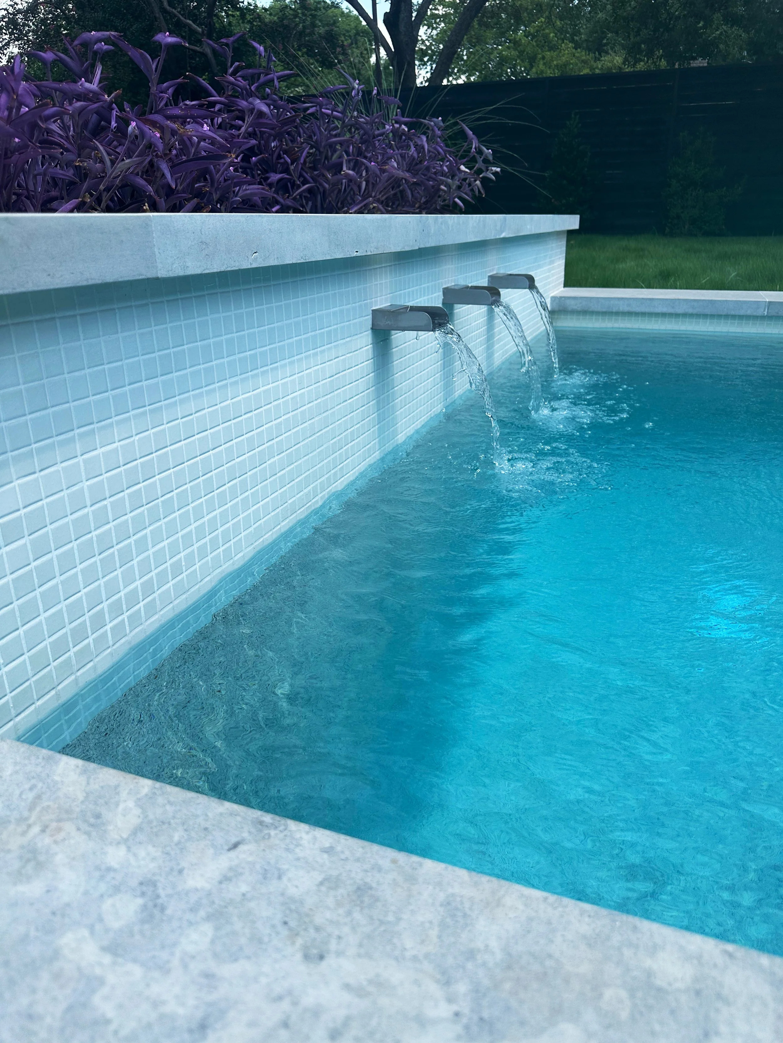 Triple Mosegi stainless steel scuppers pouring from a white glass tile wall into a modern pool