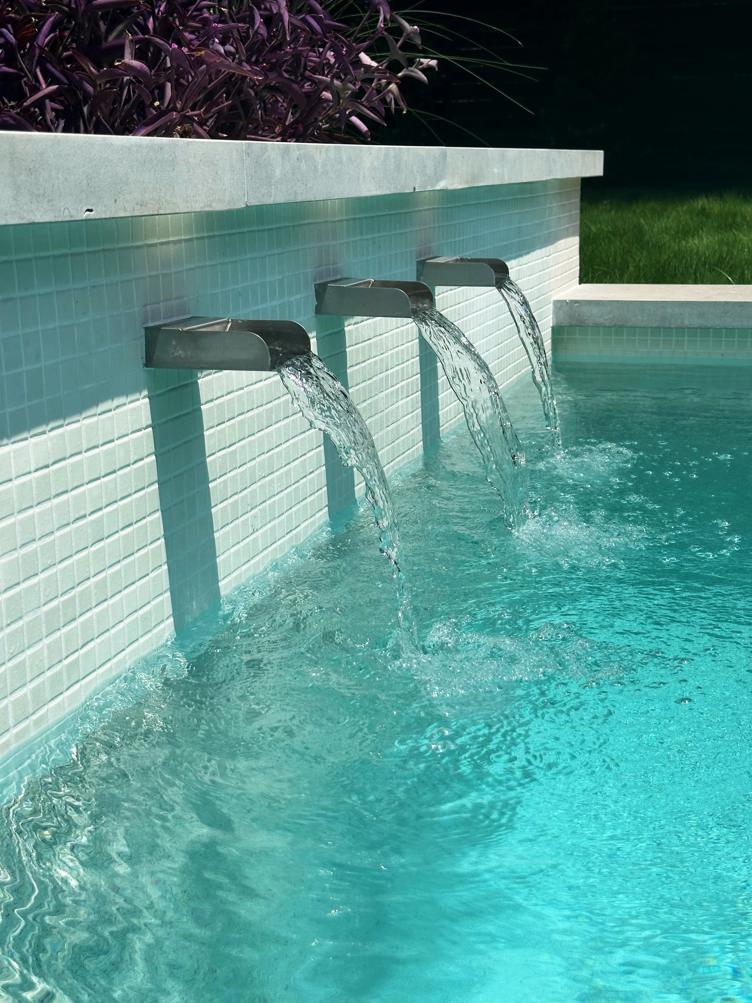 Close-up of triple Mosegi stainless steel scuppers pouring from a white tile wall topped with charcoal Leuders stone