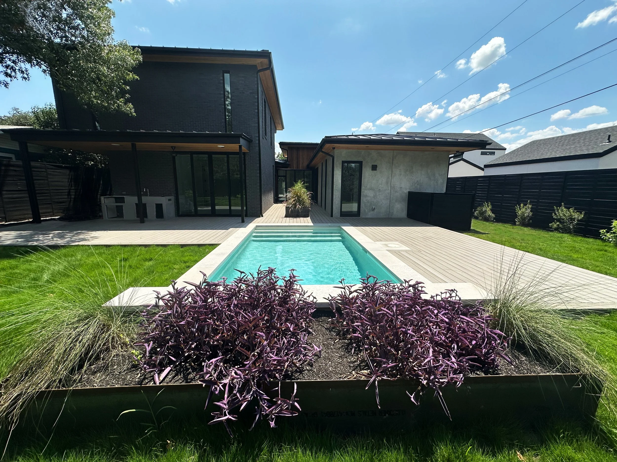 Modern pool with raised water feature wall and two-tone wooden deck behind a contemporary black brick home