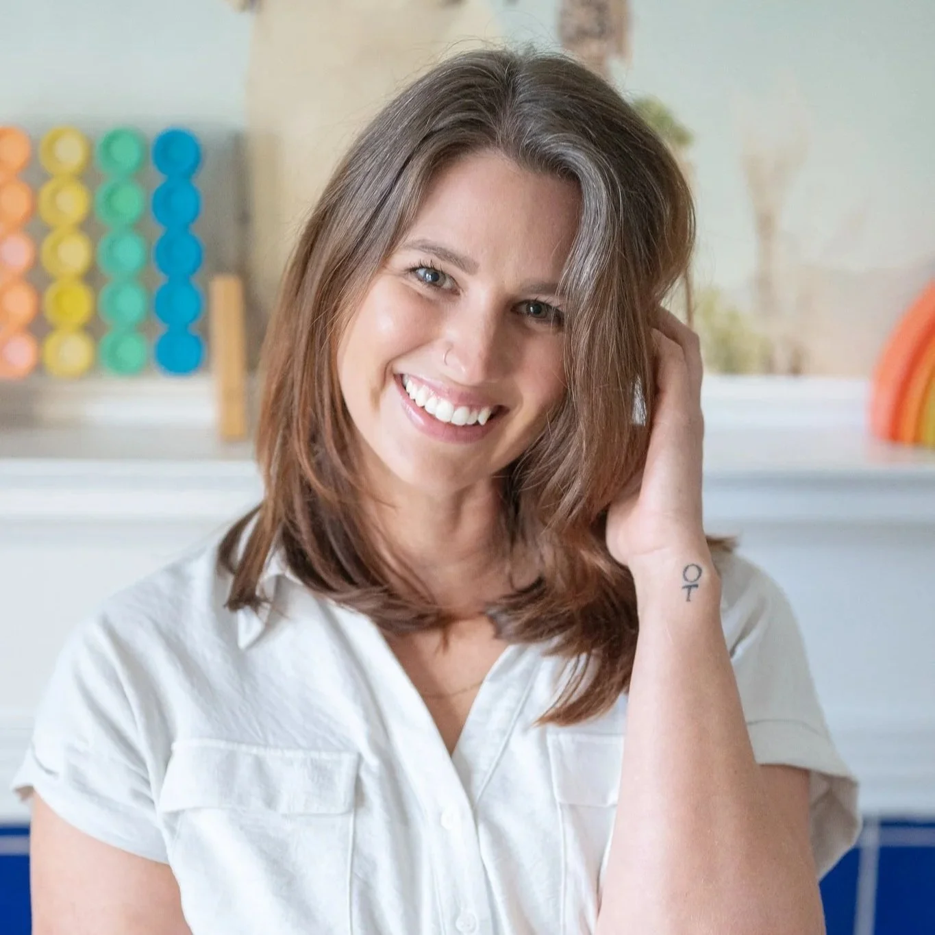 Katie Lietz, MOT, OTR/L with shoulder-length brown hair smiling, wearing a white shirt, in a colorful, playful environment with abacus and rainbow decorations in the background.