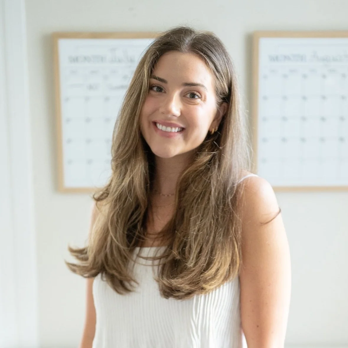 Abby Casaccia with long, wavy brown hair, wearing a white sleeveless top, standing indoors in front of whiteboards.