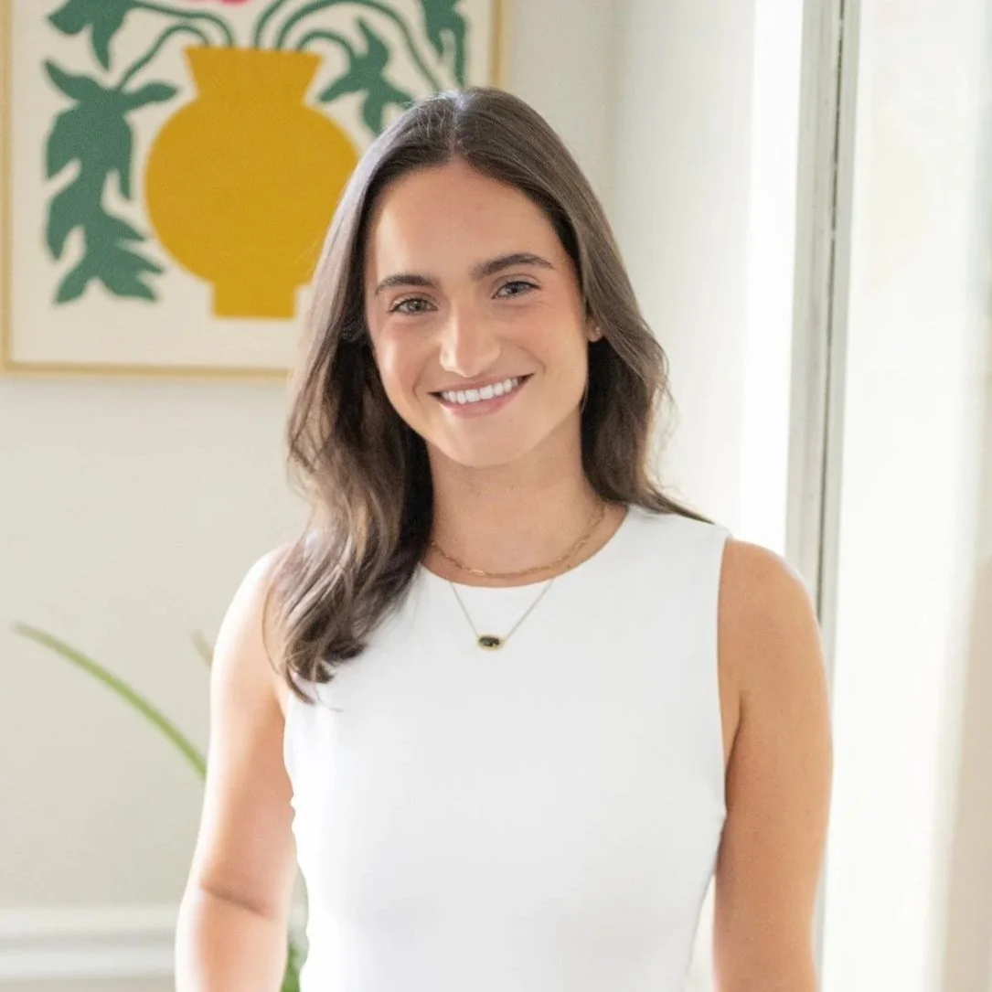 Hannah with long brown hair and a white sleeveless top smiling indoors, with a framed abstract artwork and a plant in the background.