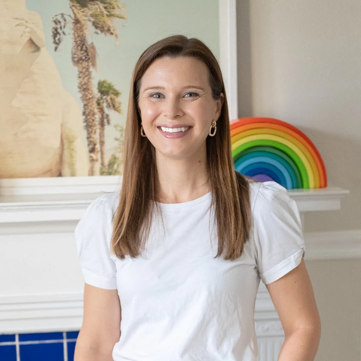 Erin Ellis with long brown hair, wearing a white shirt and gold earrings, smiling indoors with a rainbow decoration and surfboard mural in the background.