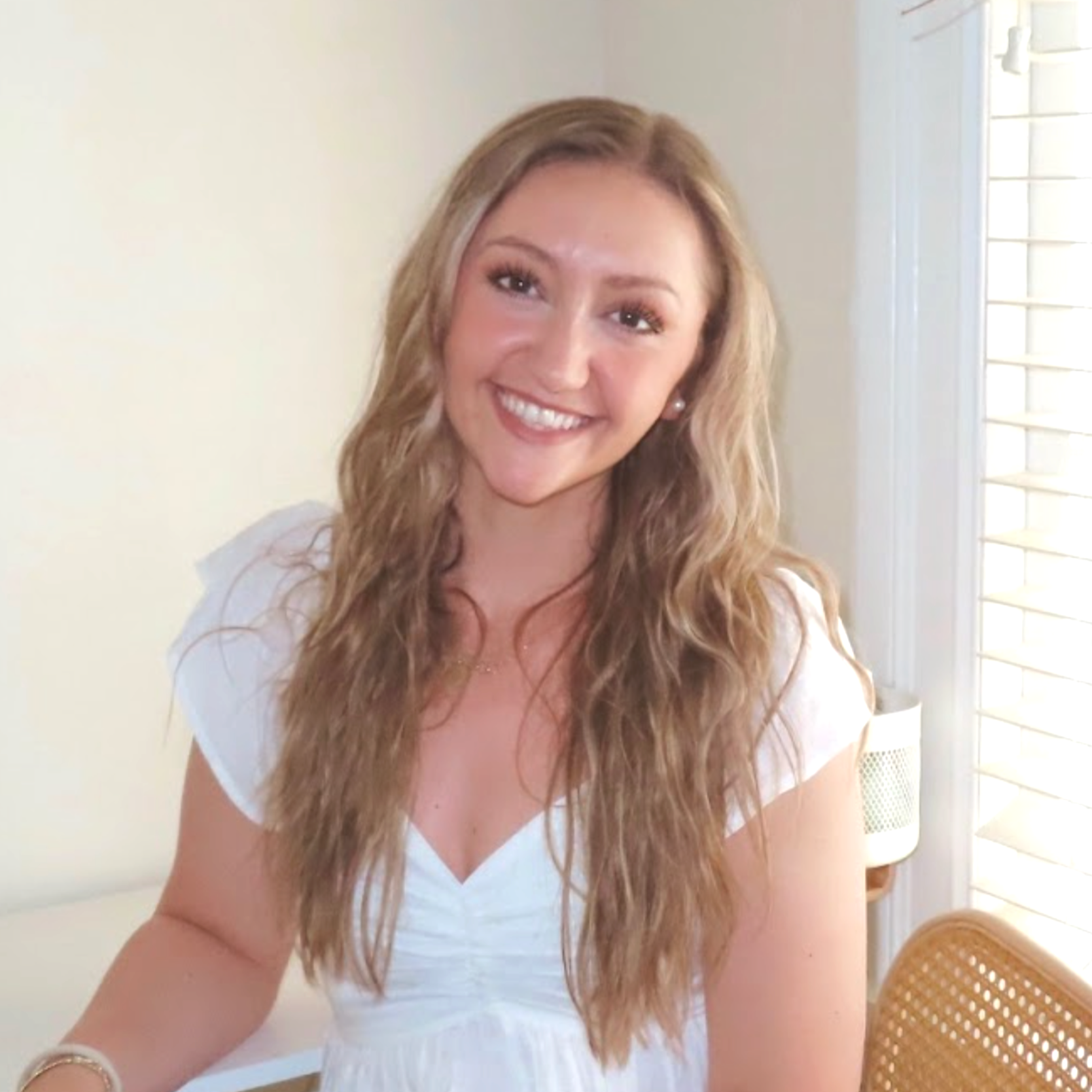 Jess Burford with long wavy blonde hair and a bright smile, wearing a white short-sleeved top, sitting indoors near a window with white blinds.